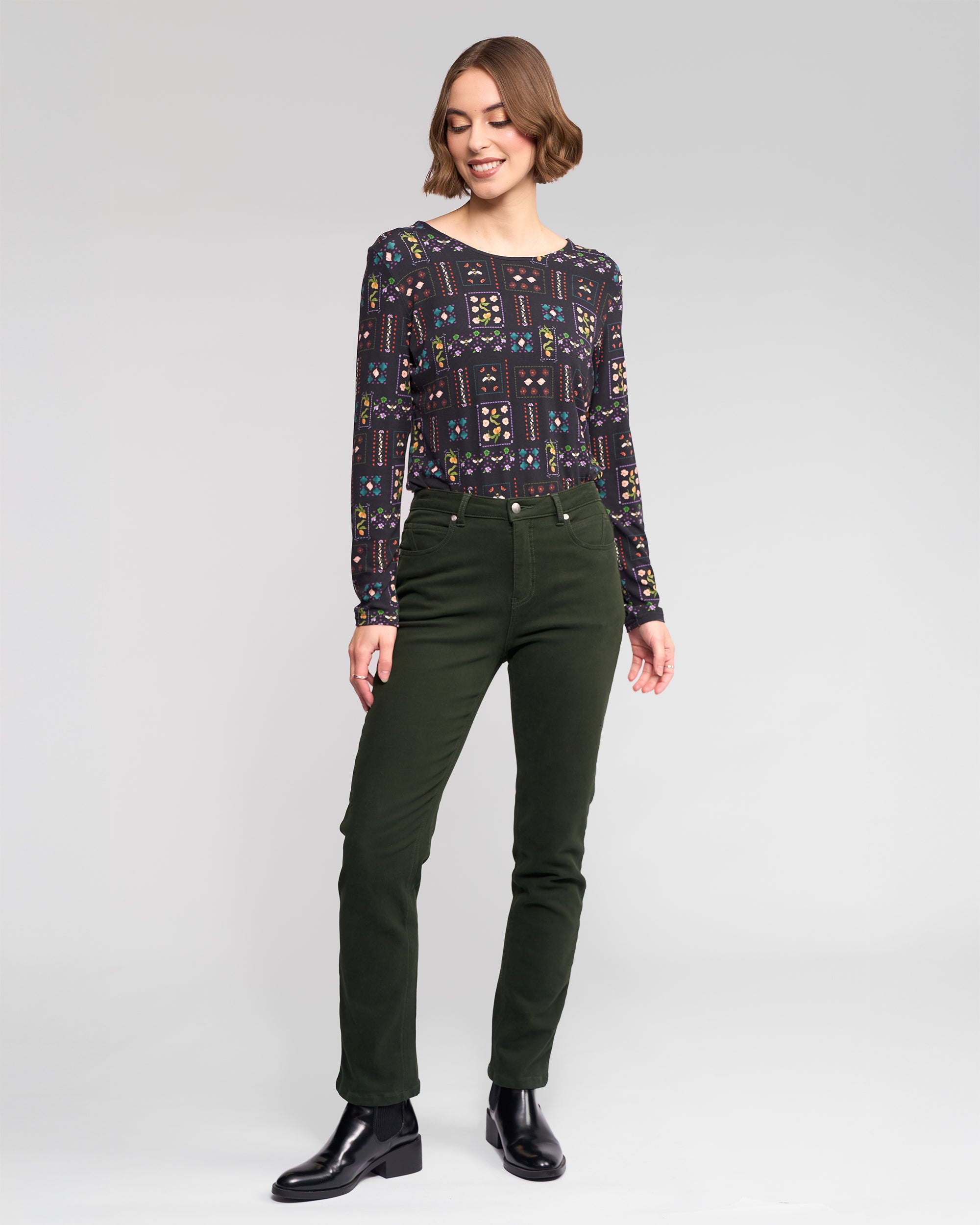 A person stands against a plain light background, wearing a patterned long-sleeve top, Vassalli Column Pant/Jean in Forest, and black ankle boots, with a slight smile and arms relaxed at their sides.