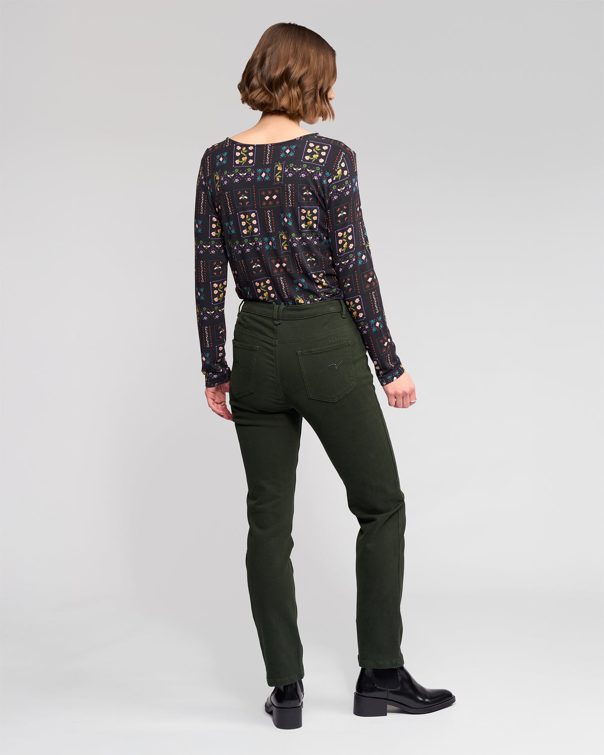 A woman stands facing away, wearing a patterned long-sleeve top, Vassalli Column Pant/Jean in Forest, and black ankle boots against a plain light gray background.