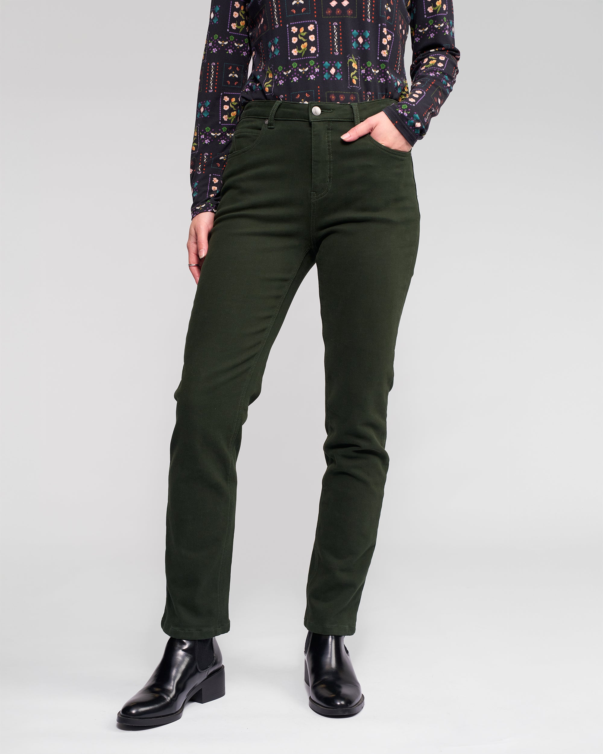 A person in Vassalli's Column Pant/Jean – Forest, a patterned long-sleeve shirt, and black ankle boots stands with one hand in their pocket against a plain light gray background.