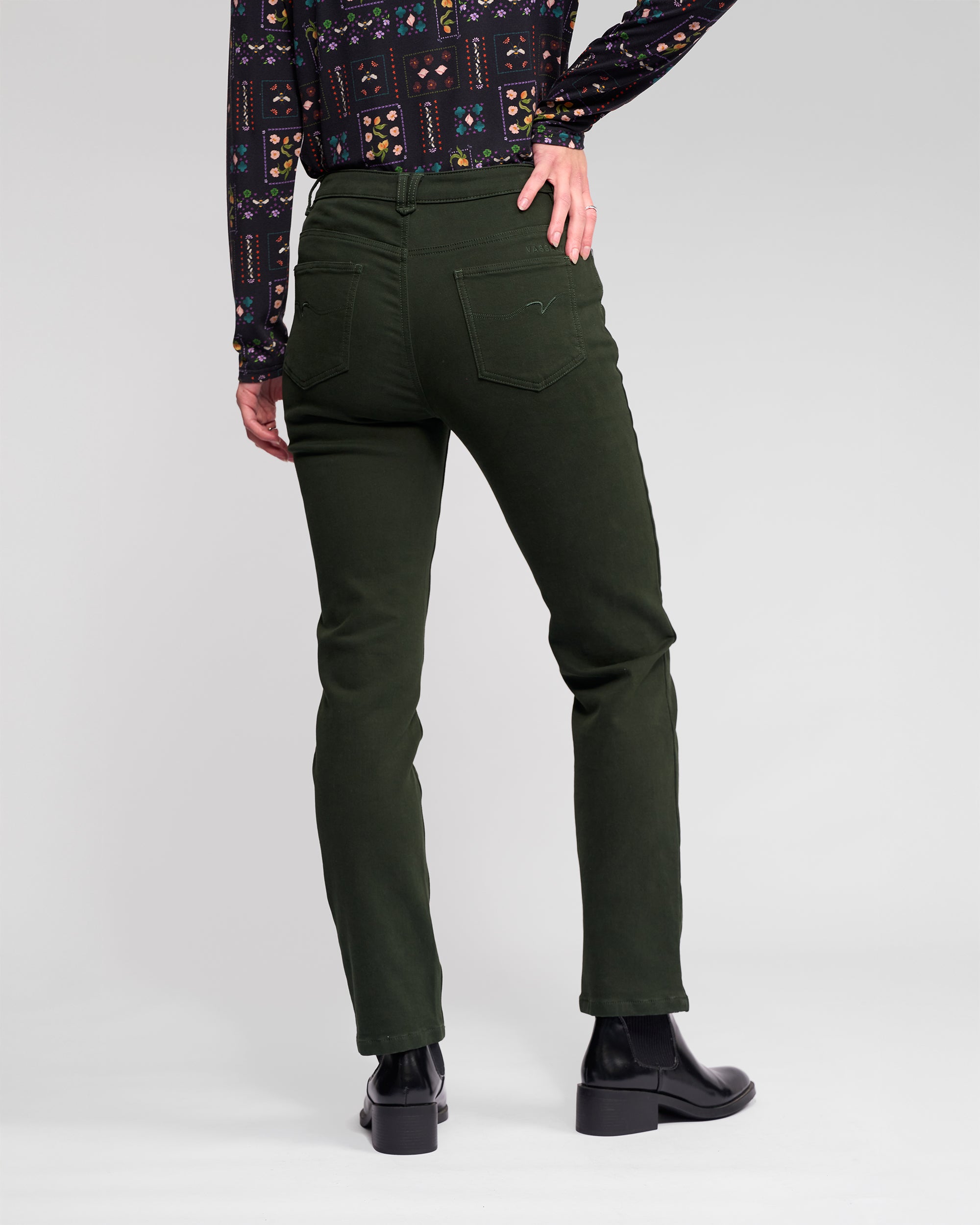 A person stands with their back to the camera, wearing Vassalli's Column Pant/Jean in Forest, a patterned long-sleeve shirt, and black ankle boots against a plain light gray backdrop.