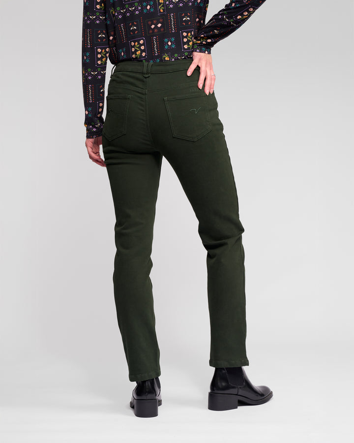 A person stands with their back to the camera, wearing Vassalli's Column Pant/Jean in Forest, a patterned long-sleeve shirt, and black ankle boots against a plain light gray backdrop.