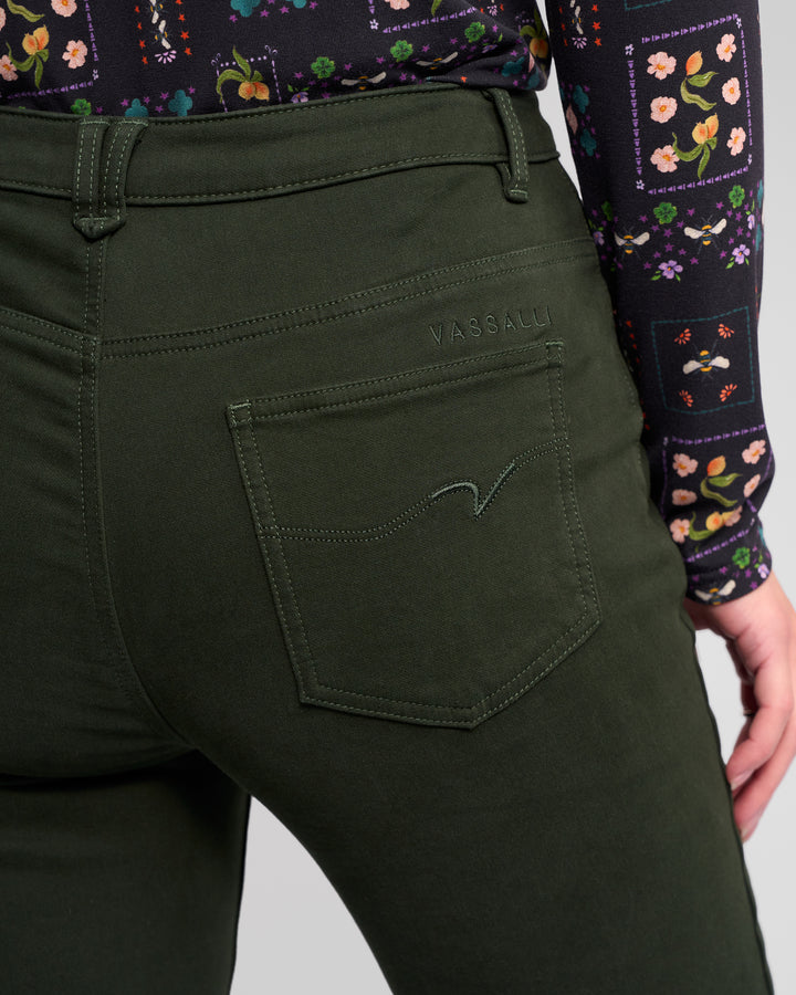 A close-up of a person wearing Vassalli Column Pant/Jean in Forest, made from stretch fabric with an embroidered back pocket, paired with a vibrant patterned long-sleeve top. Their right hand rests by their hip.