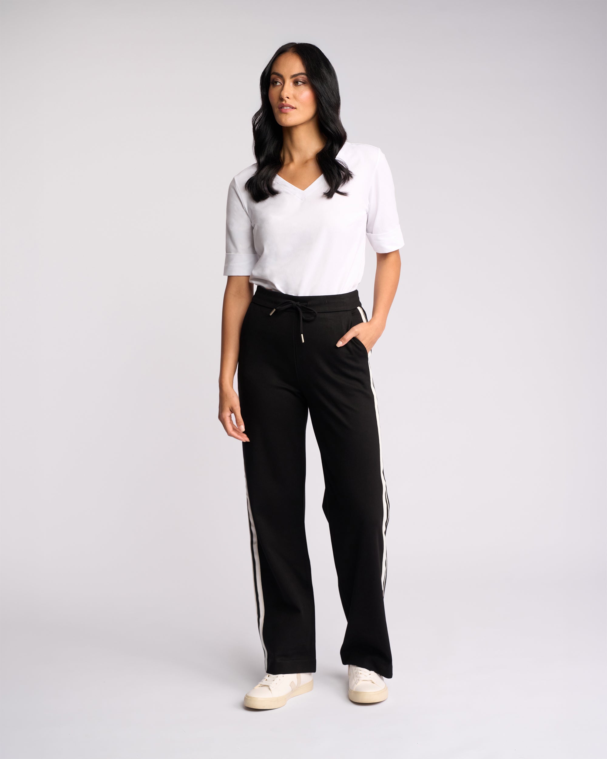 A woman with long dark hair stands against a light background wearing the Vassalli Weekend Pant - Black 2.0 with white side stripes, a white V-neck T-shirt, and white sneakers. One hand is in her pocket.