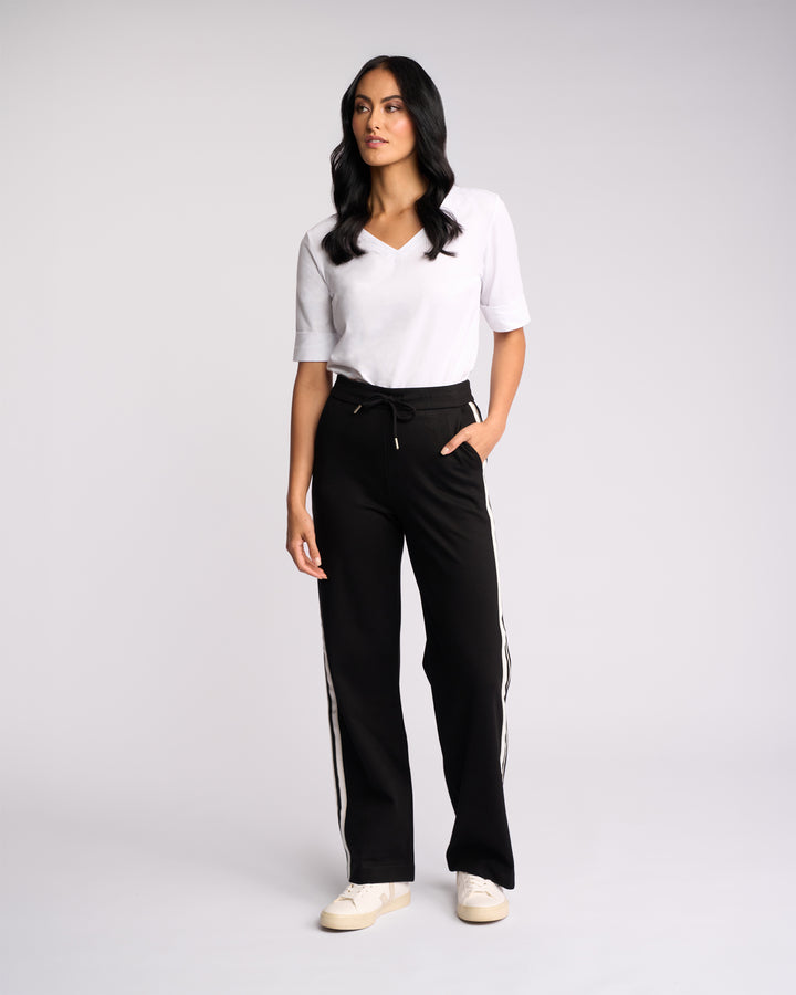 A woman with long dark hair stands against a light background wearing the Vassalli Weekend Pant - Black 2.0 with white side stripes, a white V-neck T-shirt, and white sneakers. One hand is in her pocket.