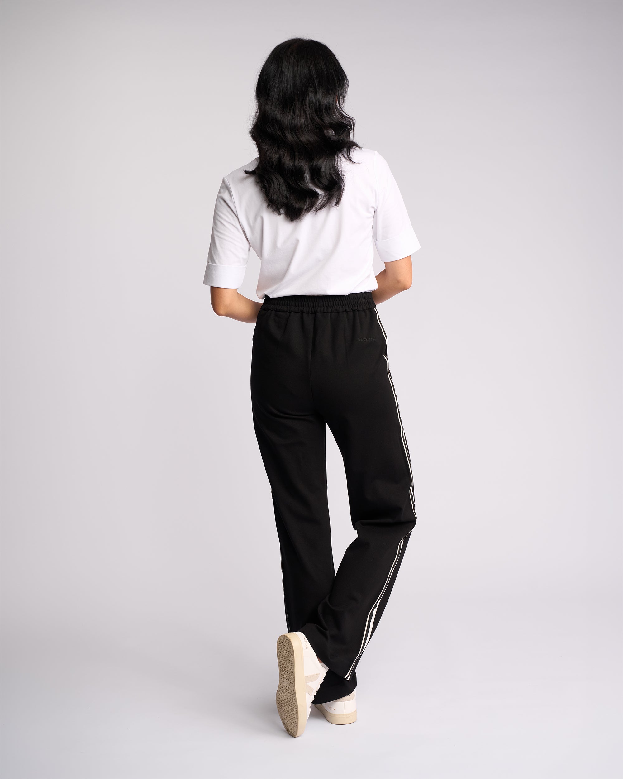 A person with long, wavy dark hair faces away, wearing a white short-sleeve shirt and Vassalli Weekend Pant - Black 2.0 with white side stripes and white sneakers, set against a plain light background.