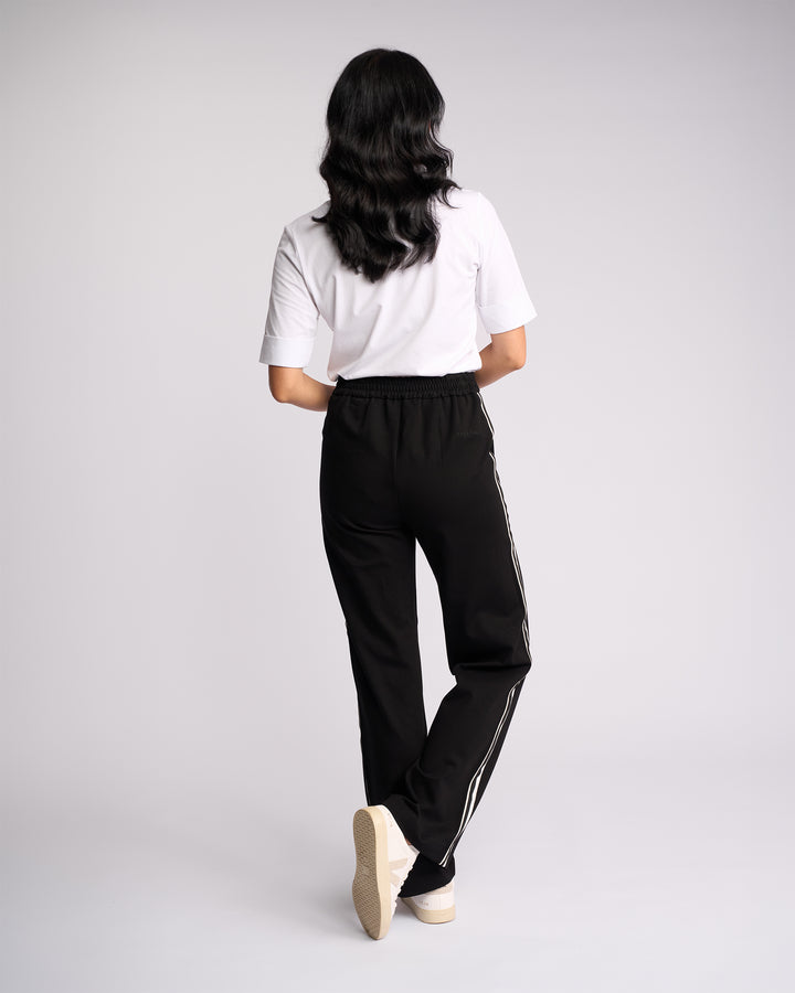 A person with long, wavy dark hair faces away, wearing a white short-sleeve shirt and Vassalli Weekend Pant - Black 2.0 with white side stripes and white sneakers, set against a plain light background.