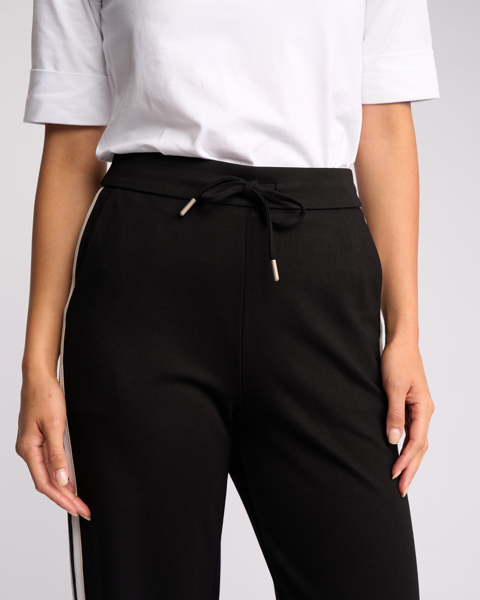 A person is shown from shoulders to mid-thigh, wearing the Vassalli Weekend Pant - Black 2.0 with white side stripes and drawstring waist, paired with a white short-sleeve top, standing against a plain background.