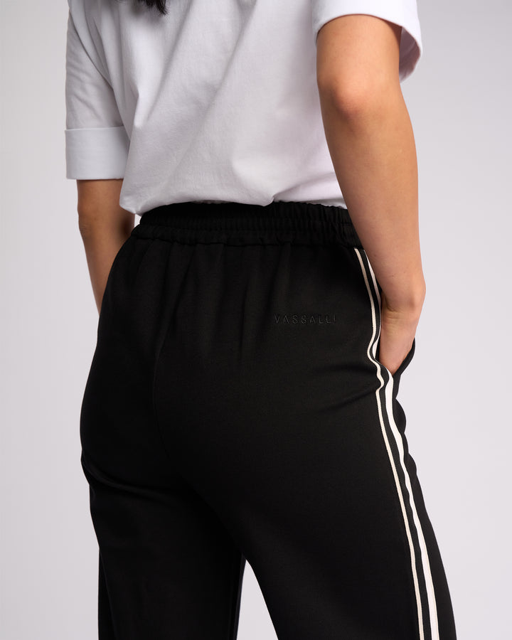 A person is seen from behind wearing a white short-sleeve shirt and Vassalli Weekend Pant - Black 2.0, which features sporty white side stripes and branded waistband, with one hand in their pocket.