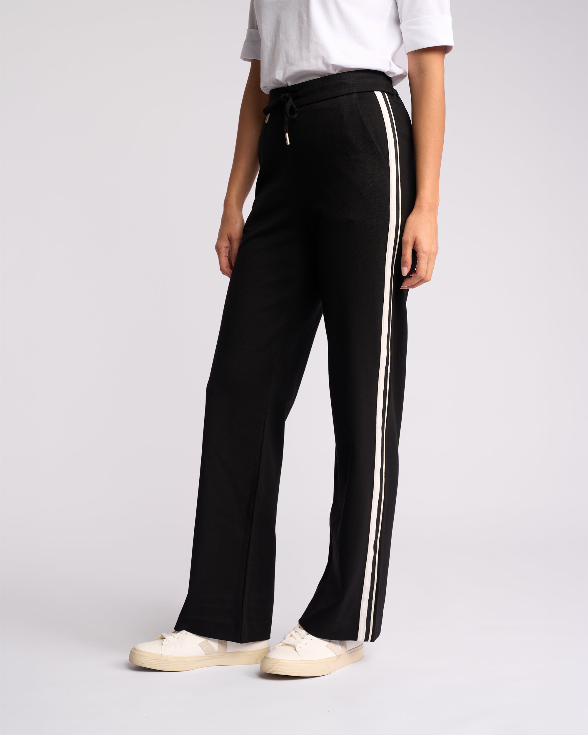 Only the lower half of a person is visible, featuring Vassalli's Weekend Pant - Black 2.0 (wide-leg with white side stripes), styled with a white short-sleeve shirt and white sneakers against a plain light background.