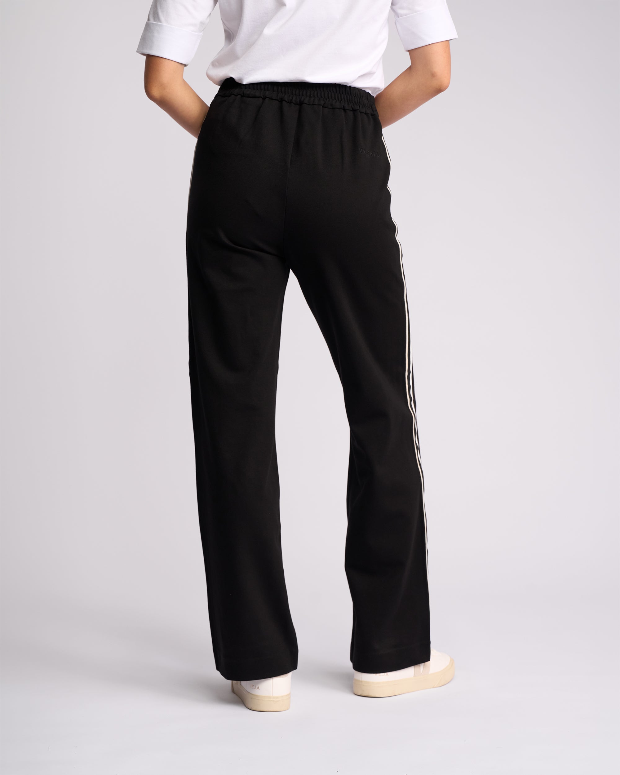 A person faces away, wearing Vassalli's Weekend Pant - Black 2.0 with white side stripes, paired with a white top and shoes, set against a plain light background.