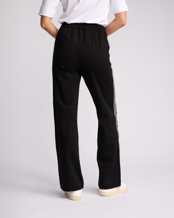 A person faces away, wearing Vassalli's Weekend Pant - Black 2.0 with white side stripes, paired with a white top and shoes, set against a plain light background.
