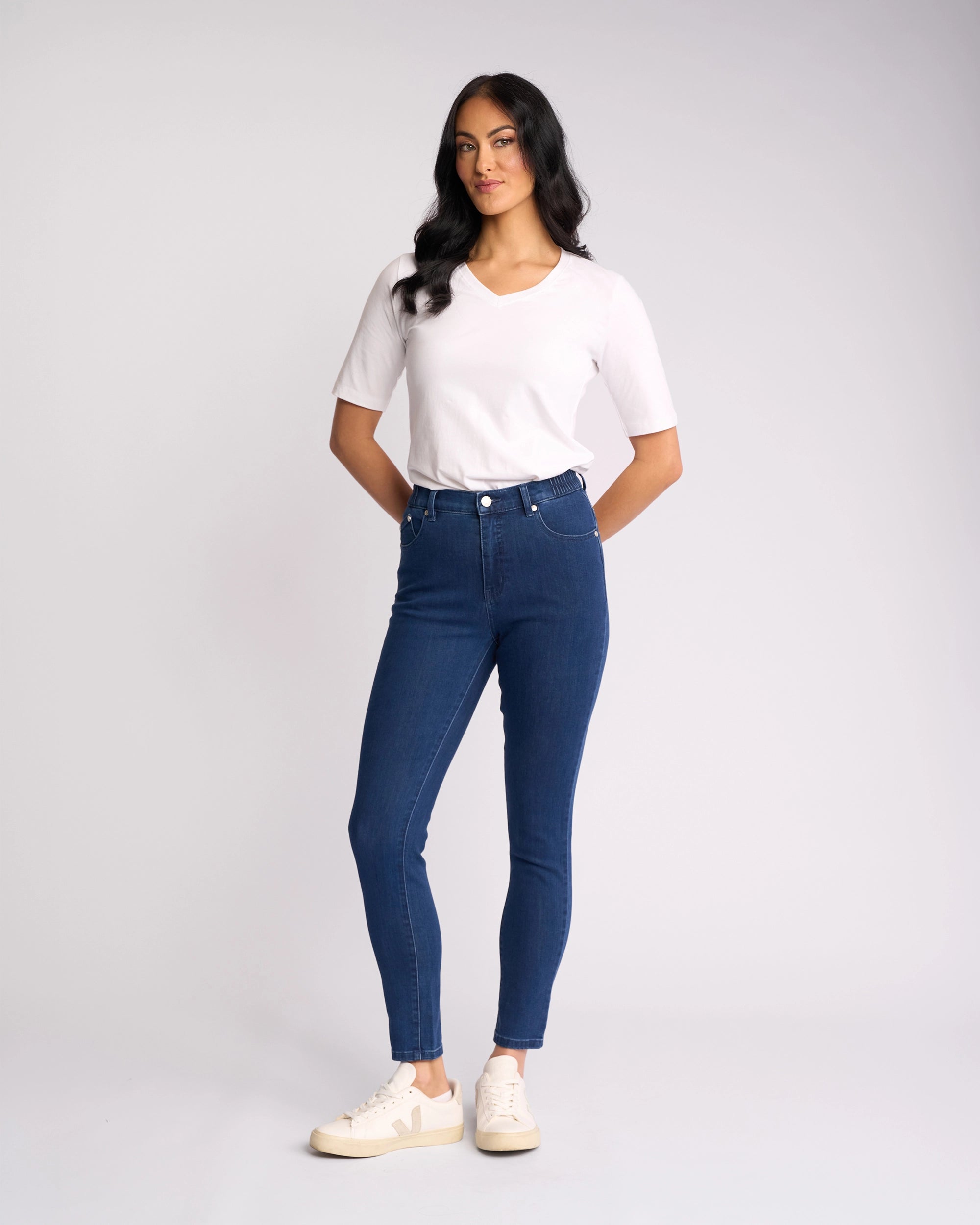 A woman with long dark hair stands against a plain background wearing a white T-shirt, white sneakers, and Vassalli’s Mode Jean - Blue Denim, featuring high-waisted skinny-leg stretch denim. Her hands are behind her back.