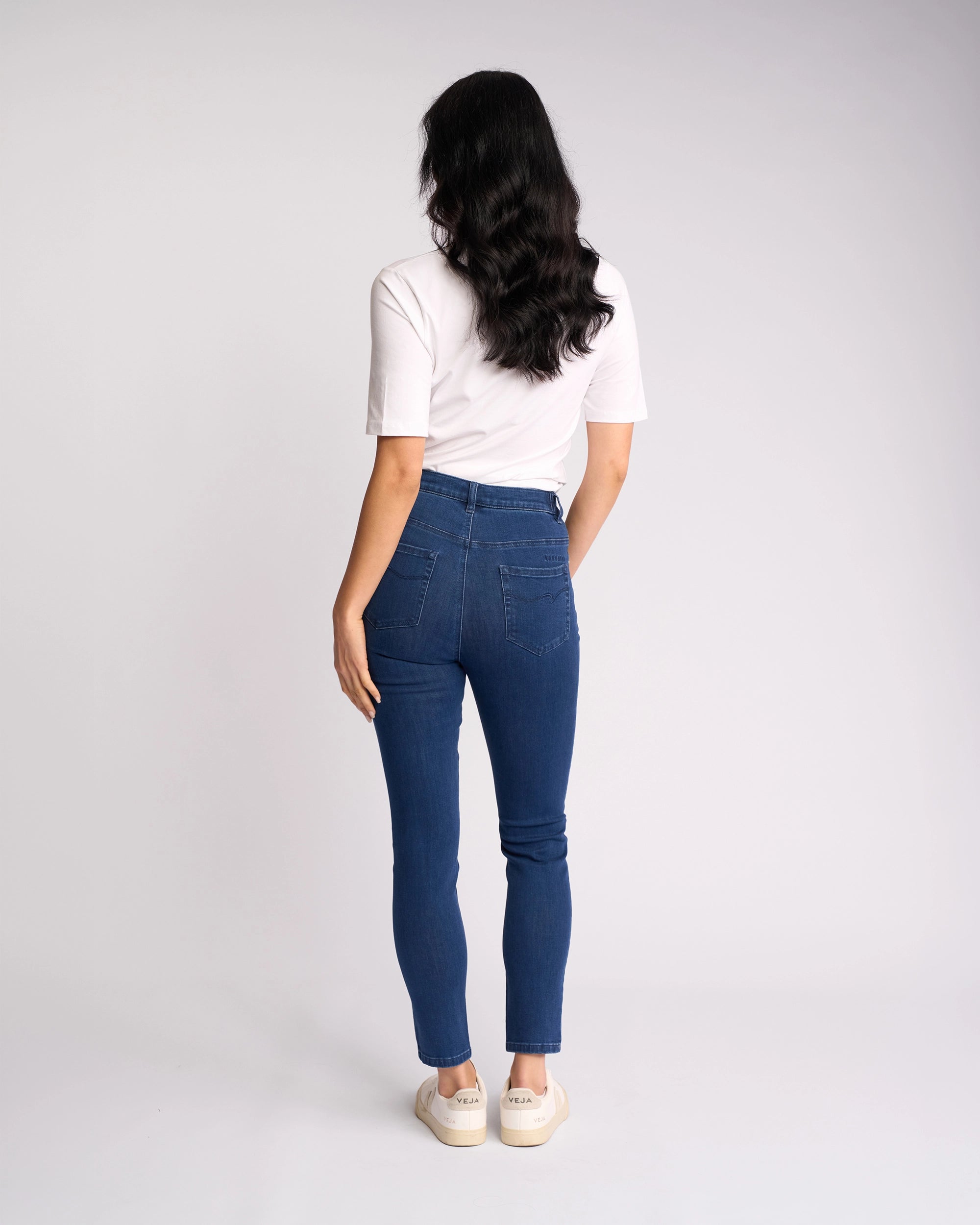A woman stands facing away in Vassalli's Mode Jean - Blue Denim, high-waisted skinny-leg jeans made from stretch denim, paired with a white t-shirt and white sneakers against a plain light background.