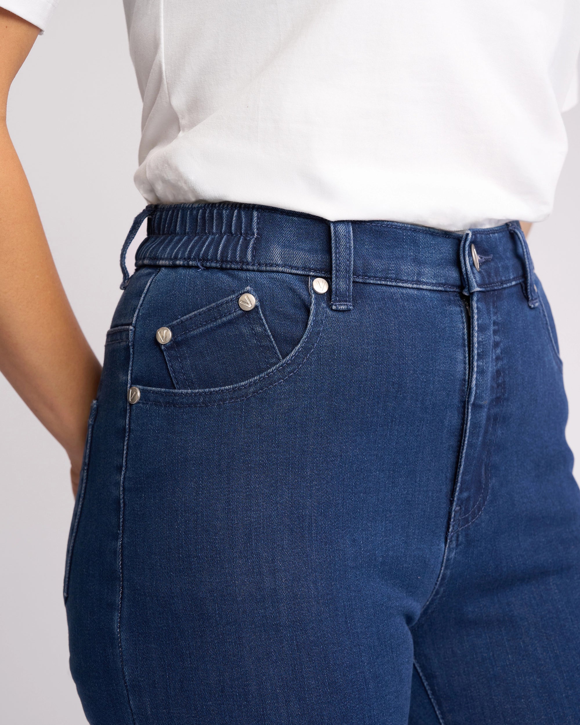 A person is shown from the waist down, slightly from the side, wearing a white shirt and Vassalli Mode Jean - Blue Denim, featuring high-waisted stretch denim with an elastic waistband.