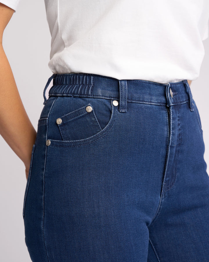 A person is shown from the waist down, slightly from the side, wearing a white shirt and Vassalli Mode Jean - Blue Denim, featuring high-waisted stretch denim with an elastic waistband.