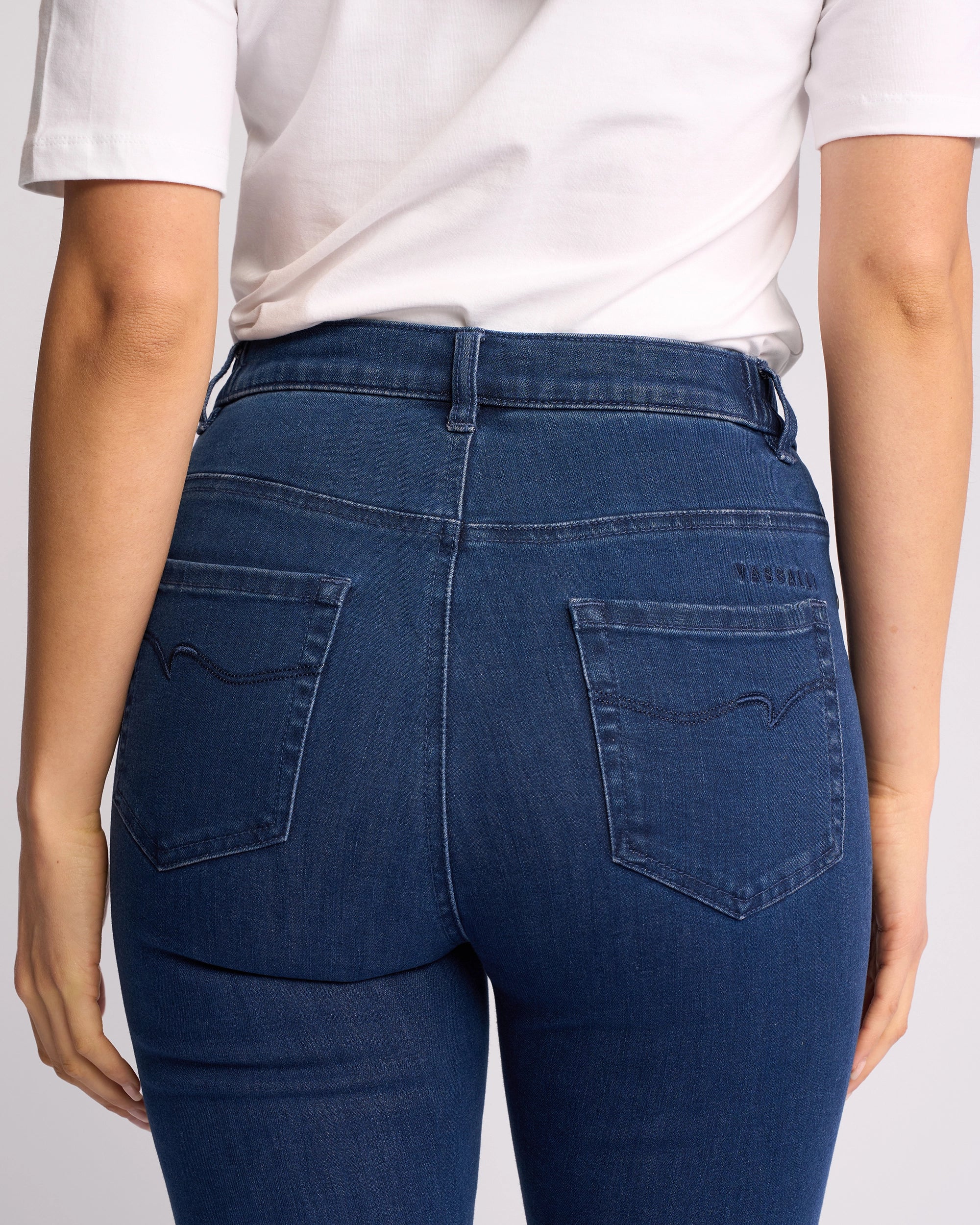A person is shown from the back wearing Vassalli's Mode Jean - Blue Denim, styled with a white t-shirt. The high-waisted, skinny-leg jeans feature two back pockets with subtle stitching and a focus on the waist and hip area.