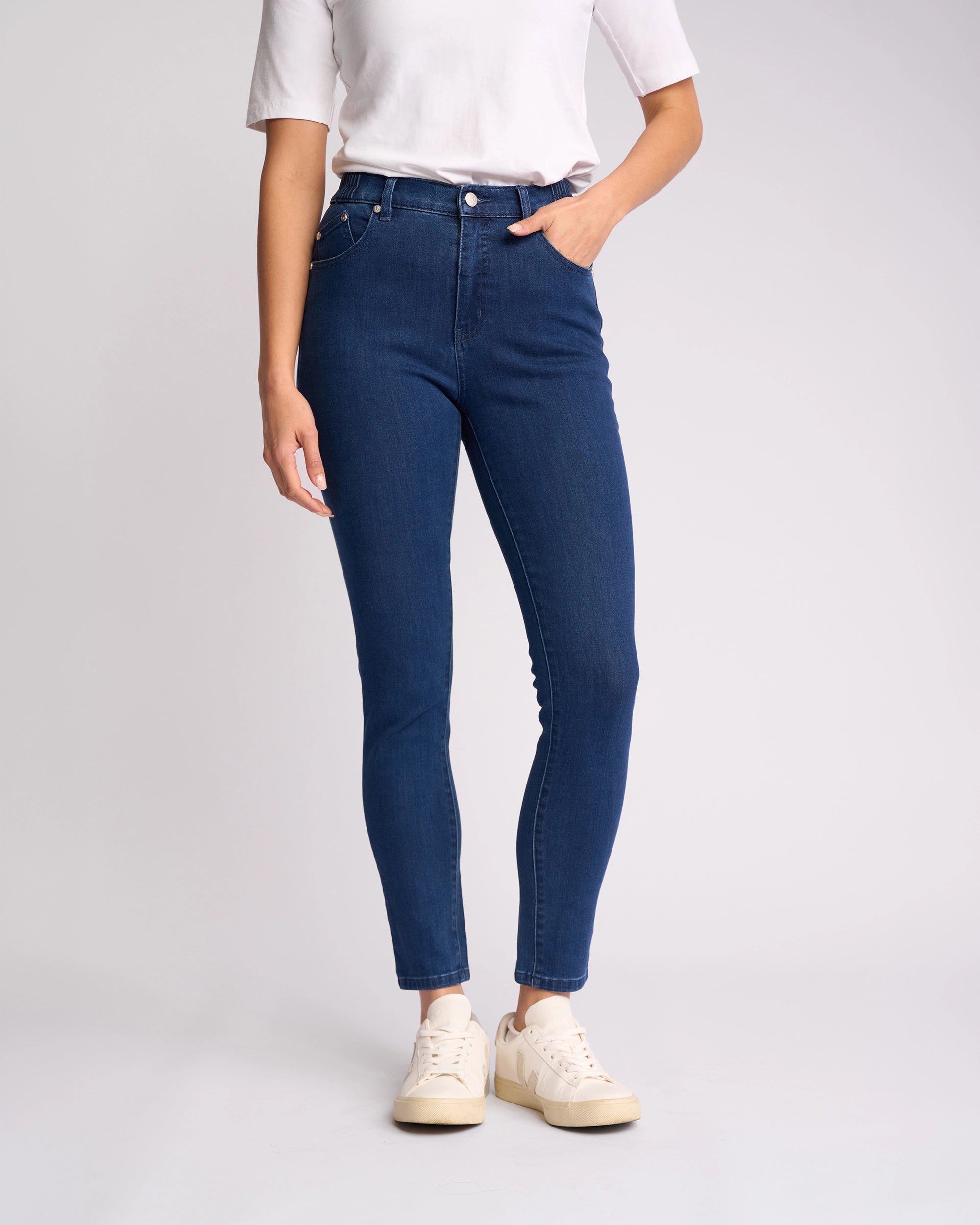 A person wears a white short-sleeve top, Vassalli Mode Jean - Blue Denim skinny-leg jeans, and beige sneakers, standing with one hand in a pocket against a plain light background.