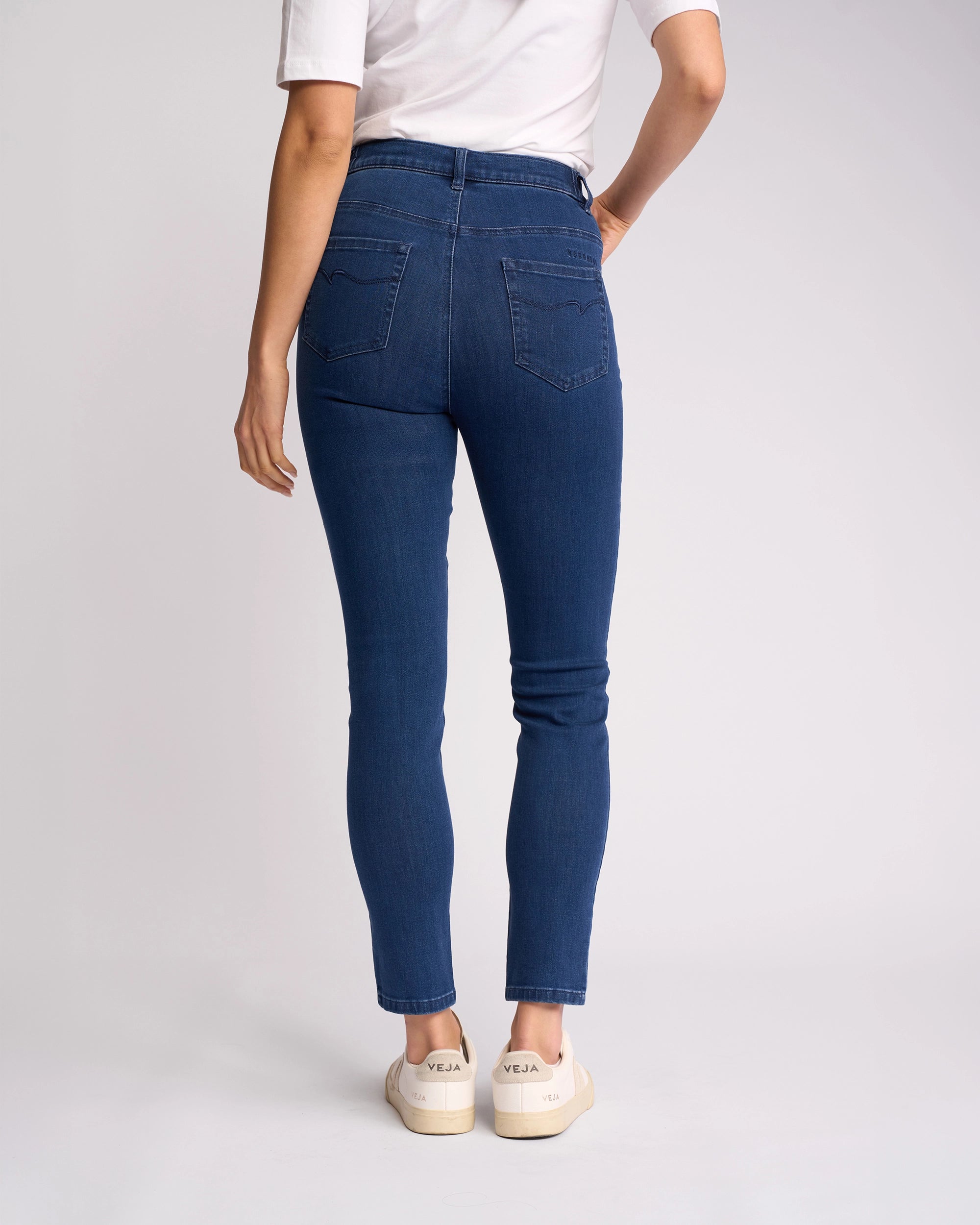 A person stands facing away, wearing a white t-shirt, Vassalli Mode Jean - Blue Denim high-waisted skinny-leg jeans made from stretch fabric, and white Veja sneakers against a plain light background.