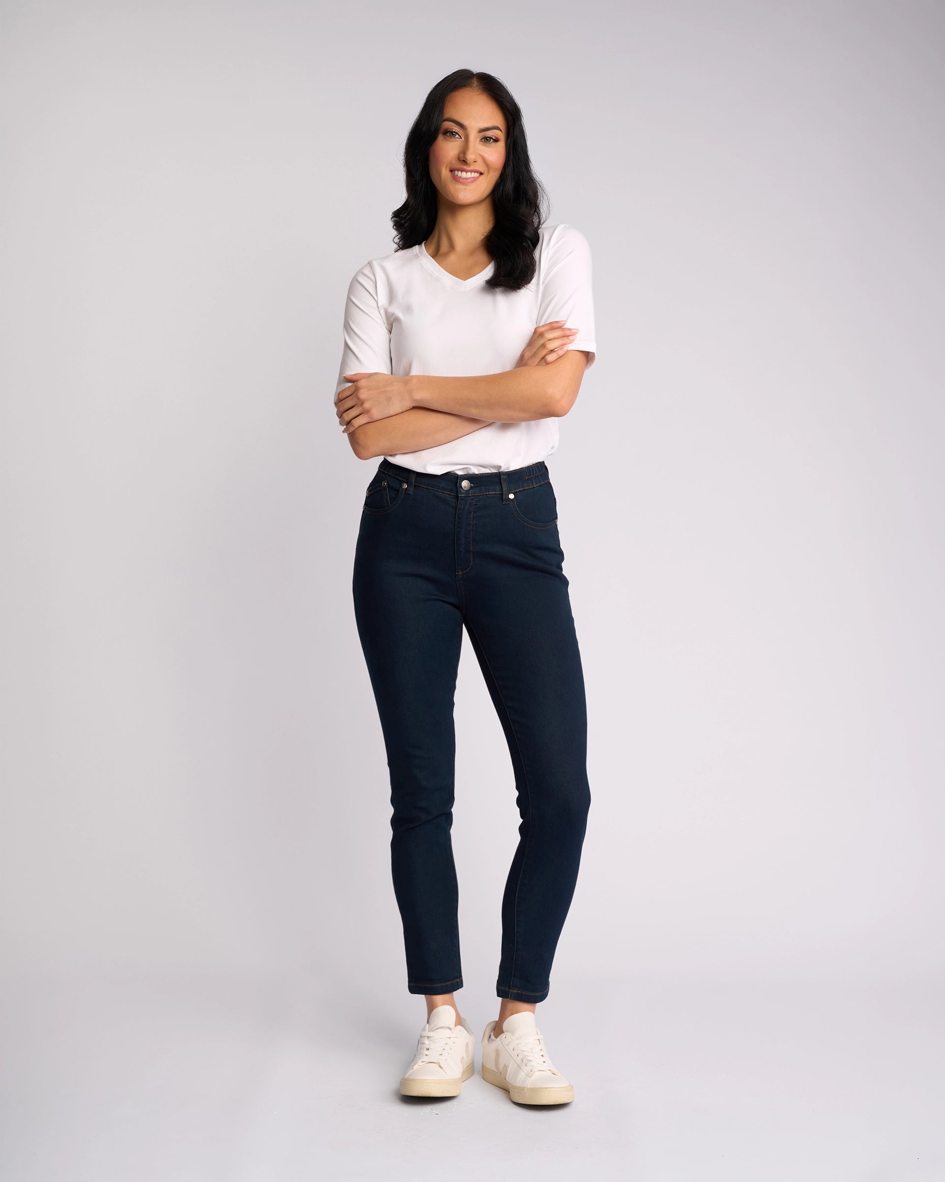 A woman with long dark hair smiles with arms crossed, wearing a white tee and Vassalli Mode Jean - Indigo Denim skinny-leg jeans made with stretch fabric, paired with white sneakers against a plain light background.