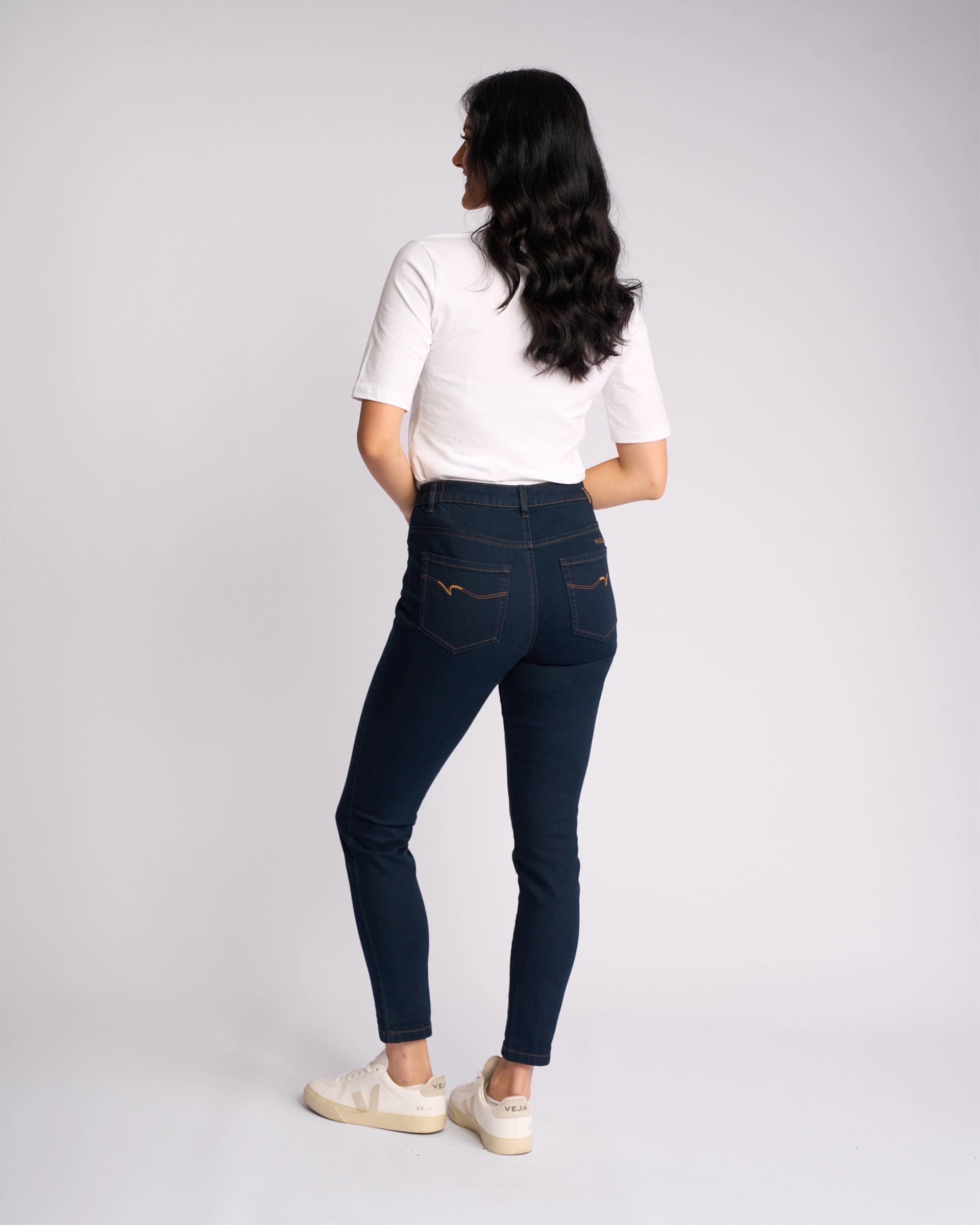 A woman faces away, wearing a white short-sleeve shirt, Vassalli Mode Jean in Indigo Denim—skinny-leg stretch jeans—and white sneakers, set against a plain light background.