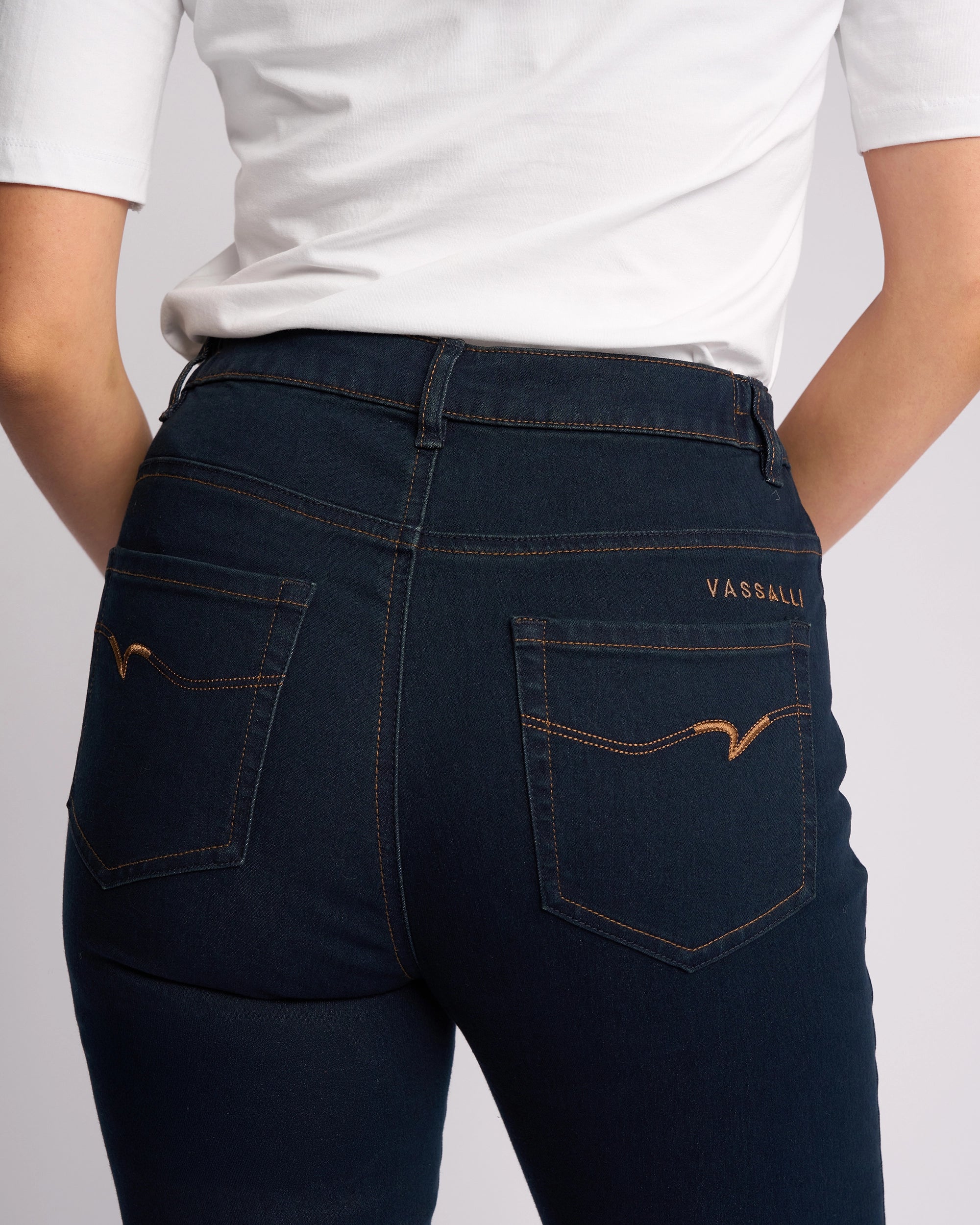 A person is shown from the back wearing a white shirt and Vassalli's Mode Jean in Indigo Denim. The skinny-leg jeans feature two back pockets with tan stitching, and they have their hands resting behind them.