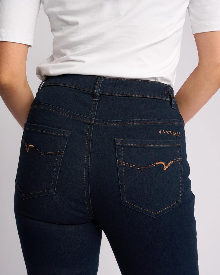 A person is shown from the back wearing a white shirt and Vassalli's Mode Jean in Indigo Denim. The skinny-leg jeans feature two back pockets with tan stitching, and they have their hands resting behind them.