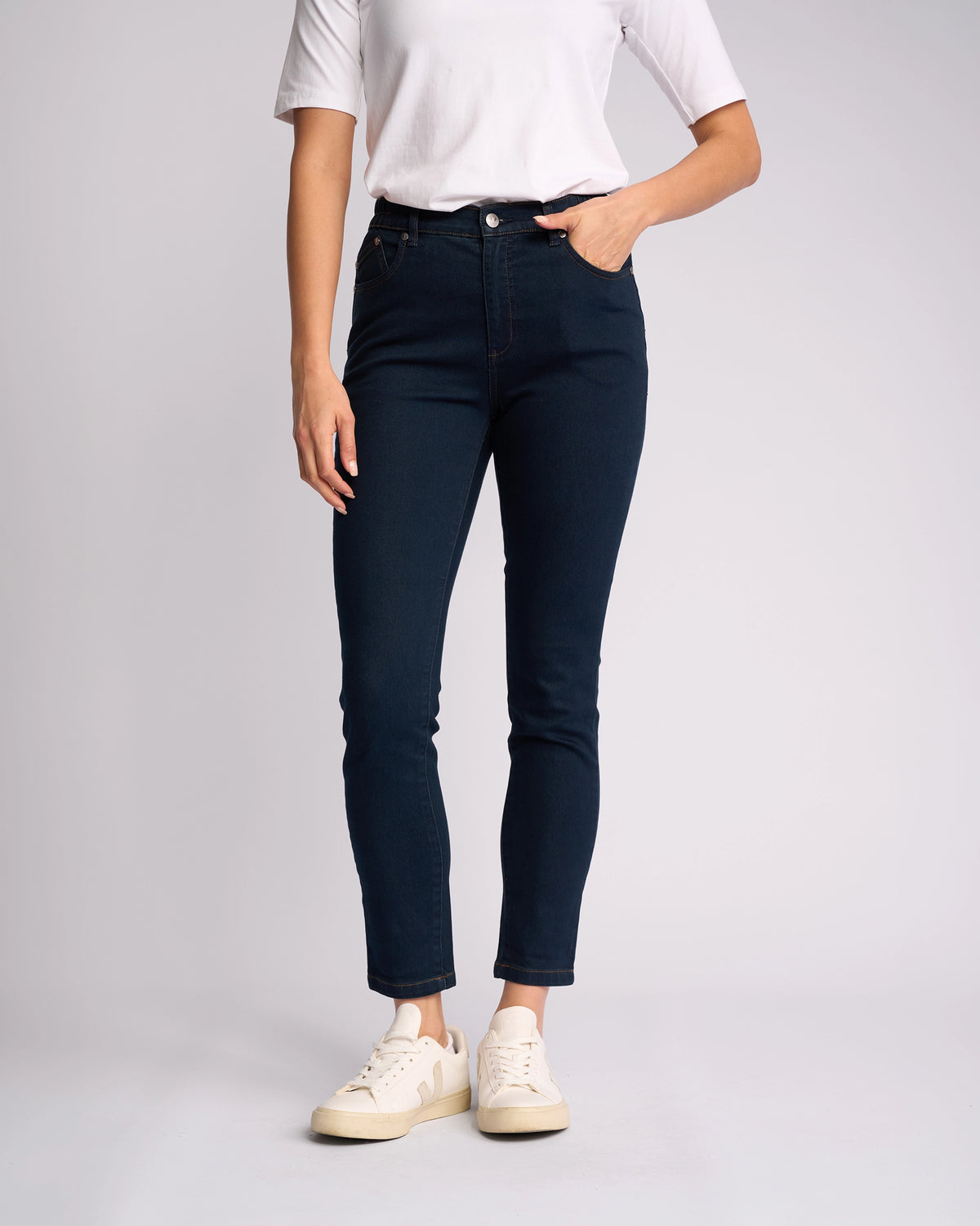 A person stands against a plain background, wearing a white short-sleeve top, Vassalli Mode Jean - Indigo Denim skinny-leg jeans made from stretch fabric, and white sneakers, with one hand in their pocket. Their face is not visible.