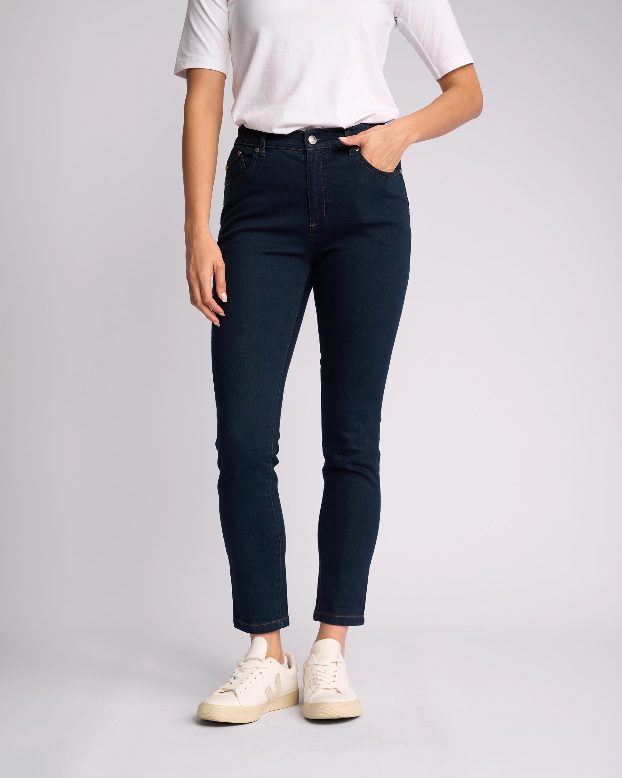 A person stands against a plain background, wearing a white short-sleeve top, Vassalli Mode Jean - Indigo Denim skinny-leg jeans made from stretch fabric, and white sneakers, with one hand in their pocket. Their face is not visible.