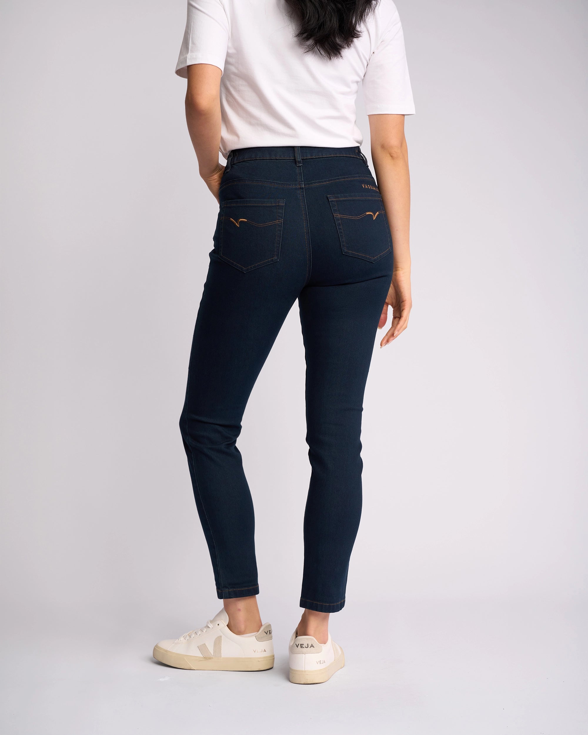 A woman faces away from the camera, wearing a white T-shirt, white sneakers with beige soles, and Vassalli Mode Jean - Indigo Denim, high-waisted skinny-leg jeans in stretch fabric, set against a plain light background.