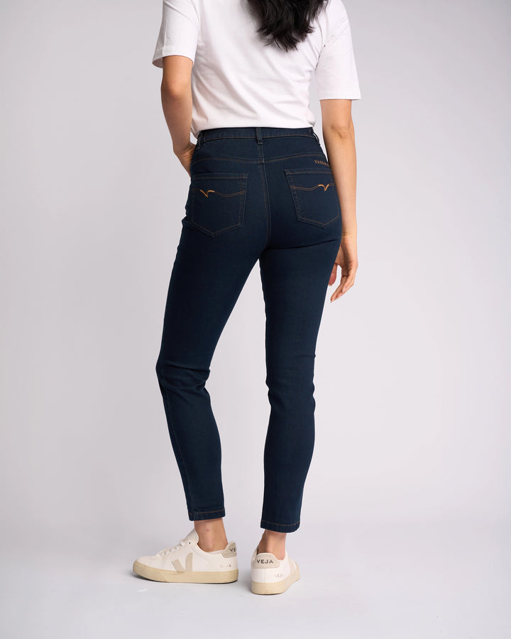 A woman faces away from the camera, wearing a white T-shirt, white sneakers with beige soles, and Vassalli Mode Jean - Indigo Denim, high-waisted skinny-leg jeans in stretch fabric, set against a plain light background.