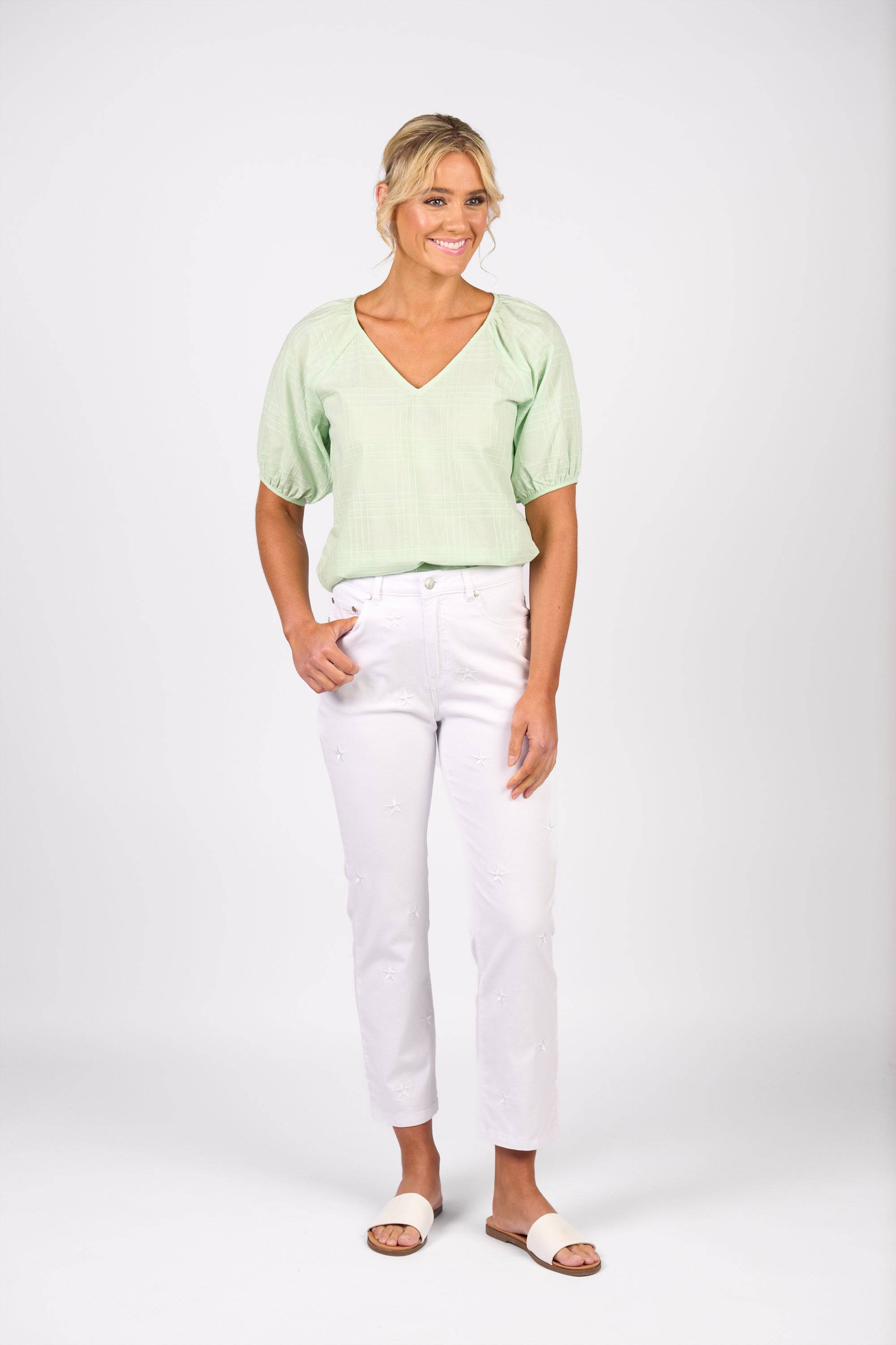 A woman smiles against a white background, wearing Vassalli's Onya Jean in White Denim with a light green short-sleeve blouse and white slide sandals. One hand is in her pocket, and her blond hair is styled in a loose updo.