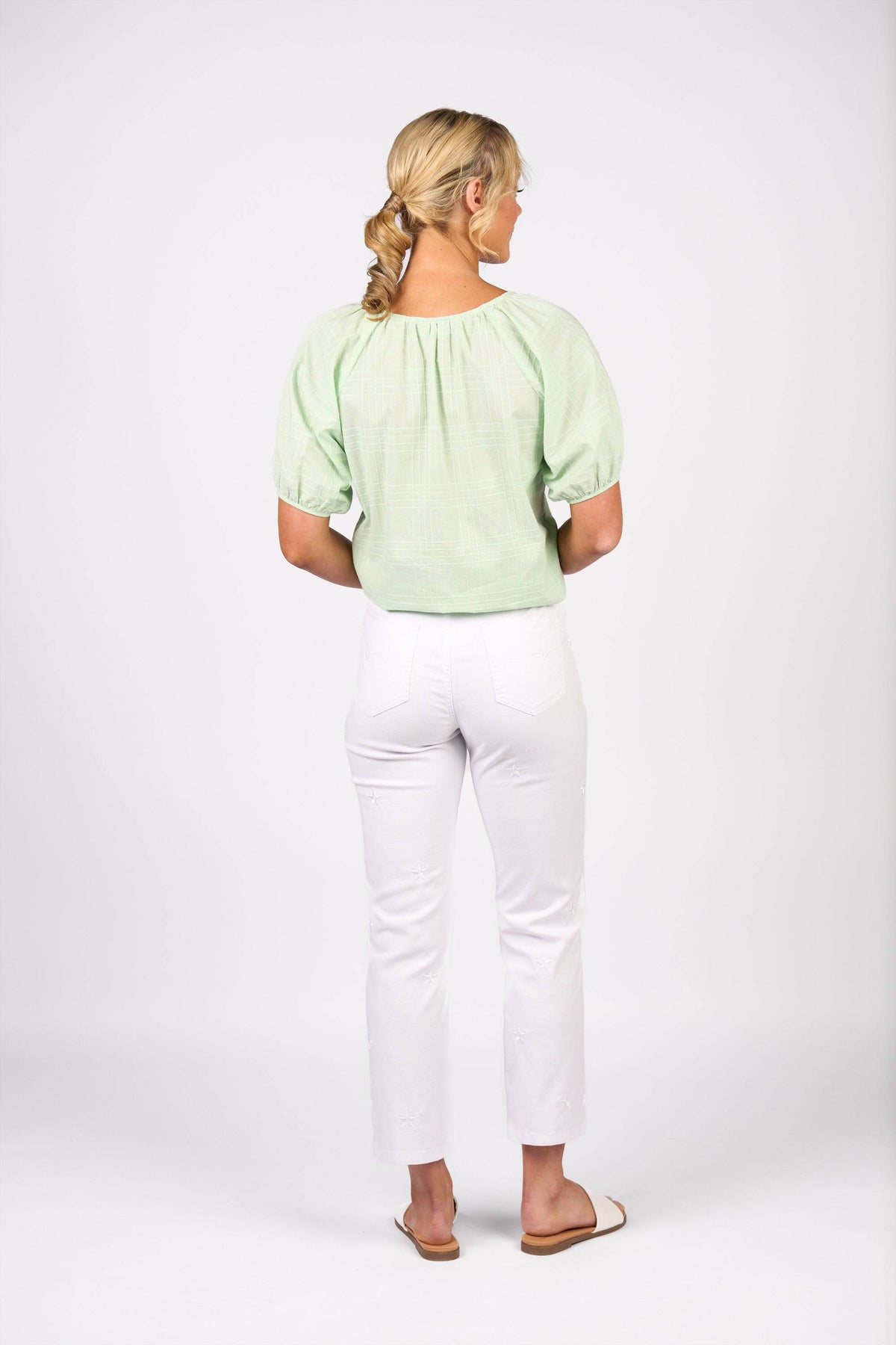 A woman faces away, wearing a light green short-sleeve blouse, Vassalli Onya Jean in white denim, and tan sandals. Her blonde hair is styled in a low ponytail against a plain white background.