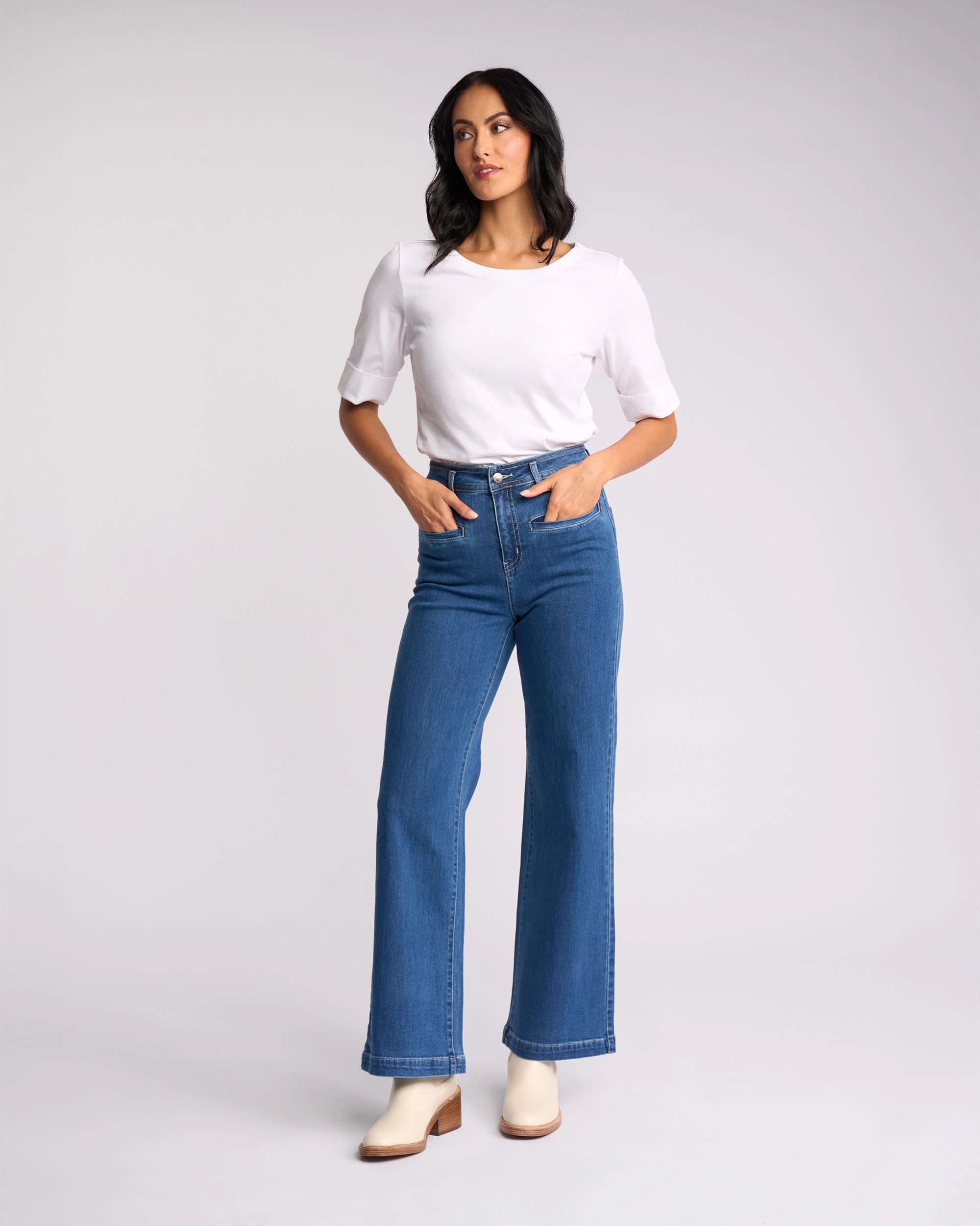 A woman with dark hair stands against a plain background, wearing the Vassalli Peace Jean in Blue Wash Denim, a white short-sleeve top, and white ankle boots. She has her hands in her pockets and looks slightly to the side.