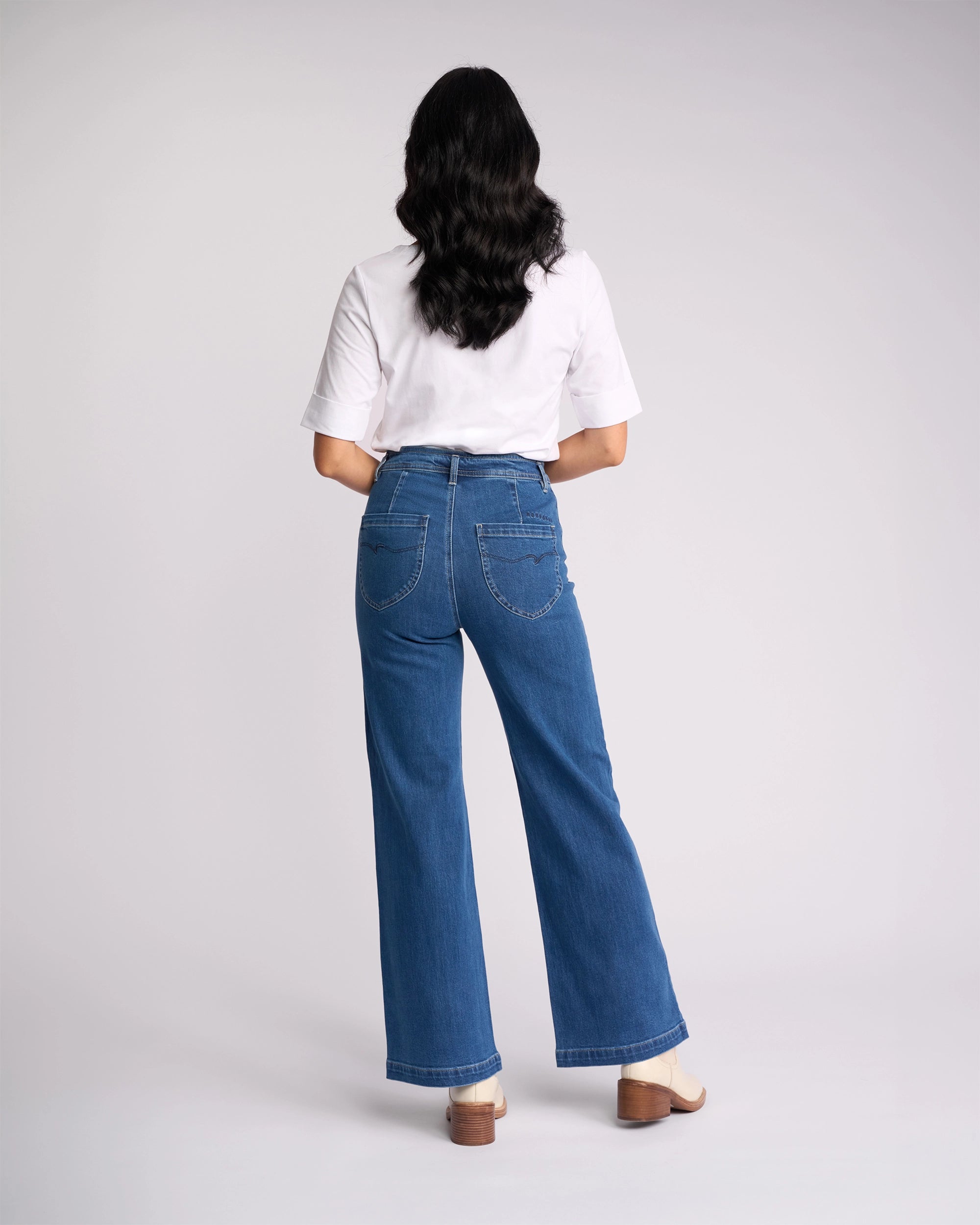 A person with long, wavy dark hair stands facing away, wearing a white short-sleeve shirt, Vassalli Peace Jean in Blue Wash Denim, and cream-colored heeled shoes against a plain light background.