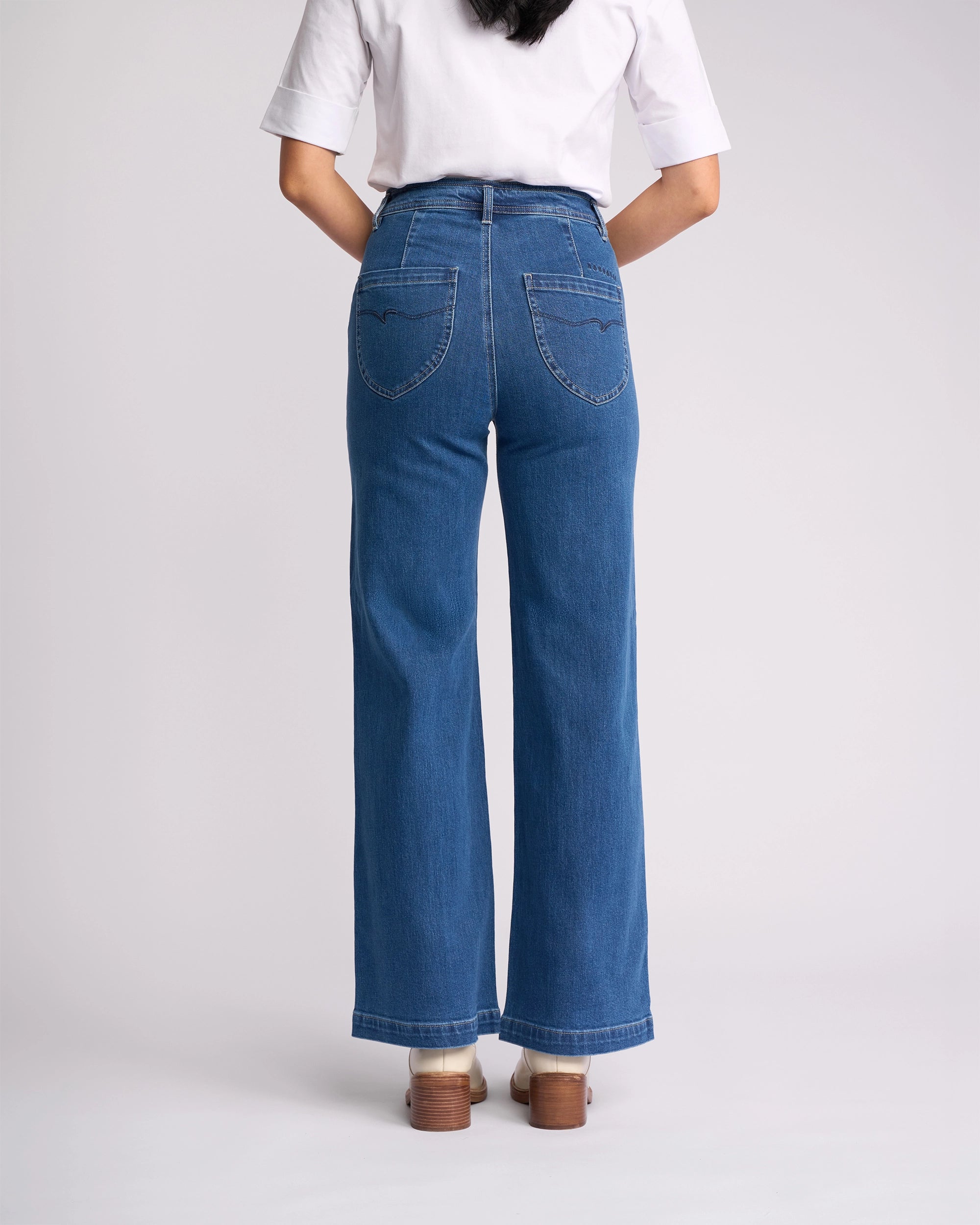 A person is seen from behind wearing a white short-sleeve shirt, brown shoes, and Vassalli's Peace Jean - Blue Wash Denim with back pockets, visible stitching, and wide, slightly flared legs reflecting the latest women’s denim NZ style.