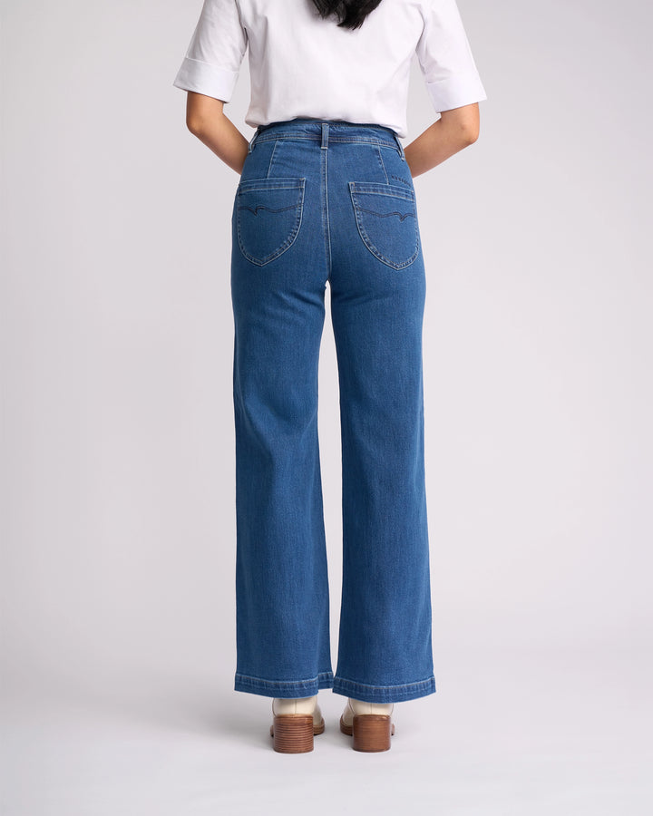 A person is seen from behind wearing a white short-sleeve shirt, brown shoes, and Vassalli's Peace Jean - Blue Wash Denim with back pockets, visible stitching, and wide, slightly flared legs reflecting the latest women’s denim NZ style.