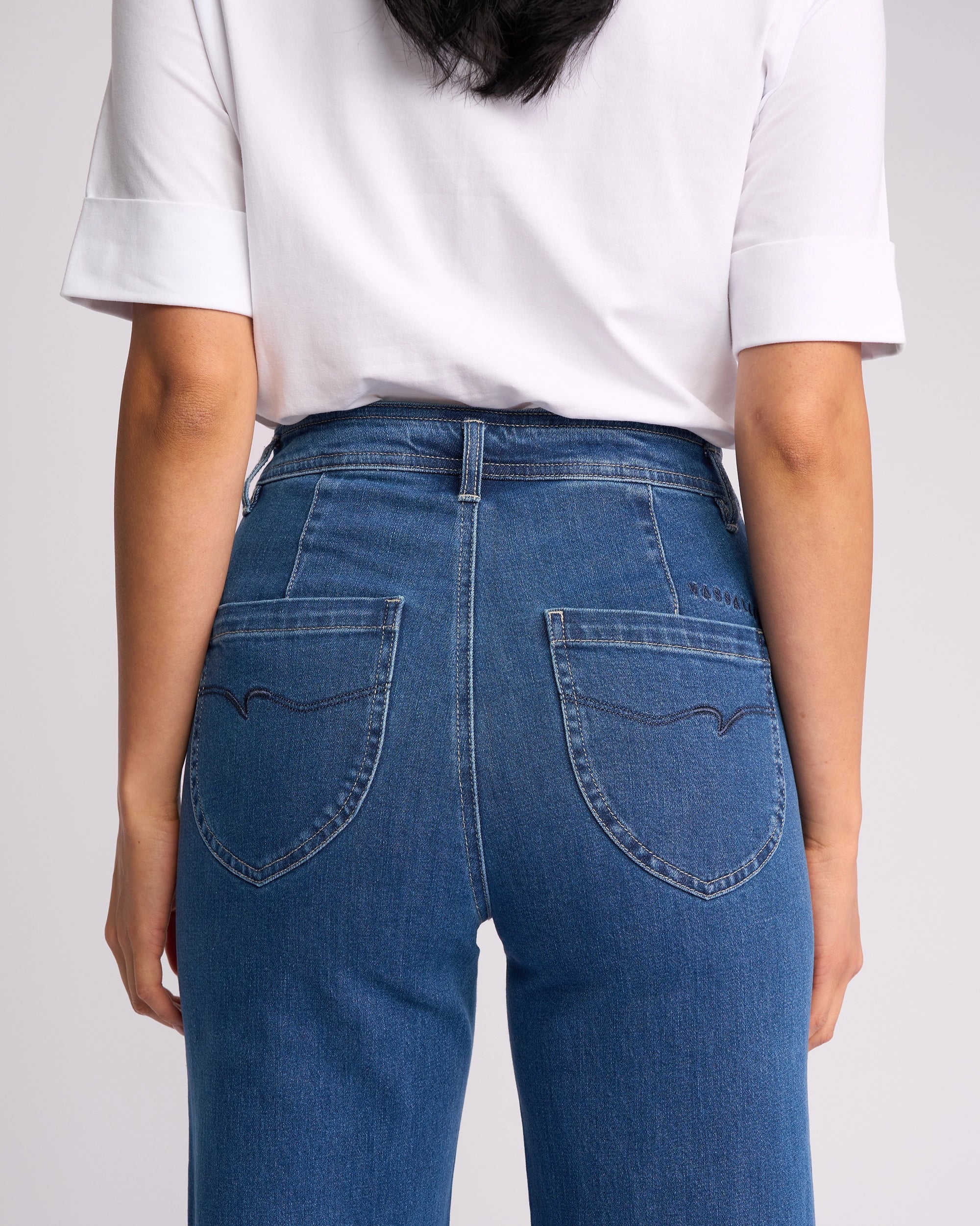 A person is seen from the back wearing a white short-sleeve shirt and Vassalli's Peace Jean in Blue Wash Denim, standing against a plain backdrop.