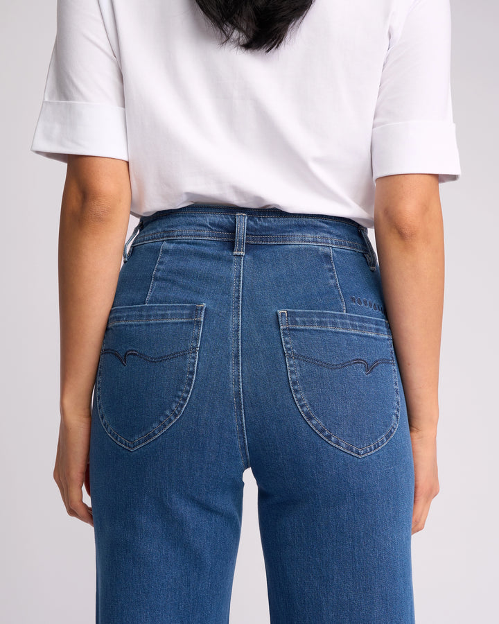 A person is seen from the back wearing a white short-sleeve shirt and Vassalli's Peace Jean in Blue Wash Denim, standing against a plain backdrop.