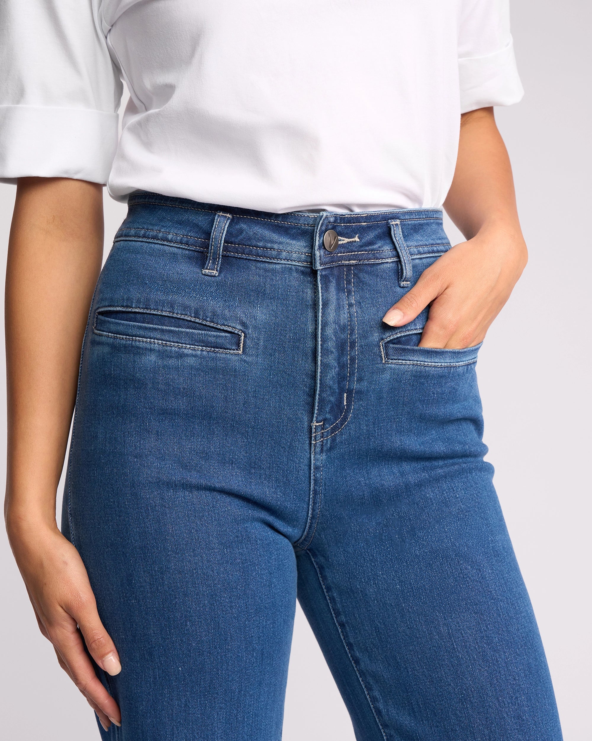 A person wears the Vassalli Peace Jean in blue wash denim, featuring front slit pockets, and a white shirt, standing with one hand in a pocket.