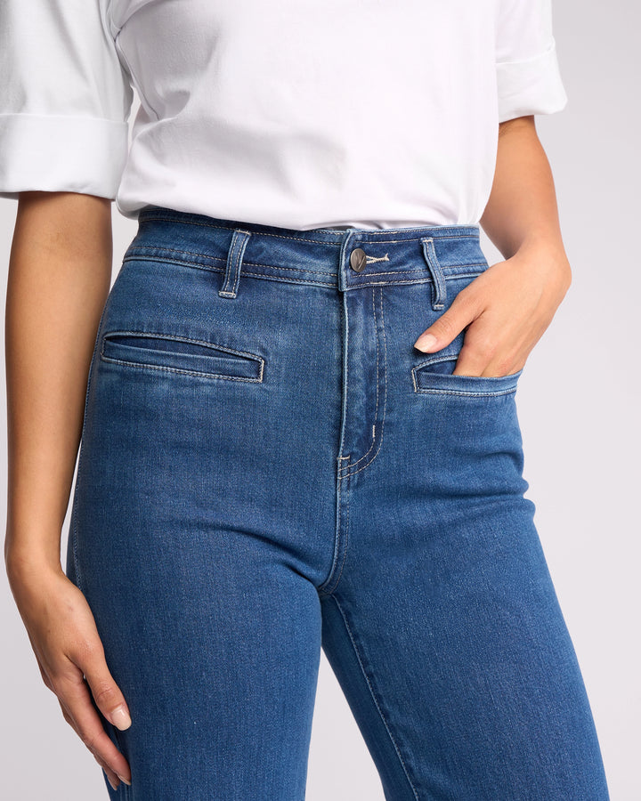 A person wears the Vassalli Peace Jean in blue wash denim, featuring front slit pockets, and a white shirt, standing with one hand in a pocket.