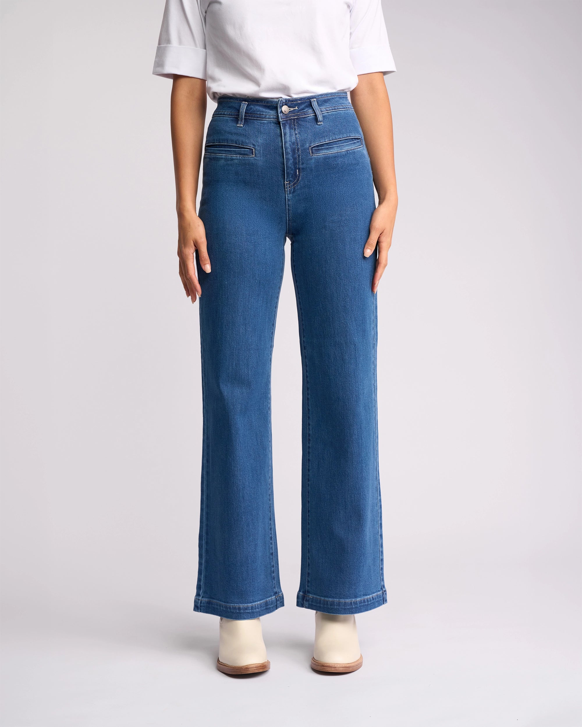 A person in Vassalli's Peace Jean - Blue Wash Denim and a white short-sleeve shirt with white heeled boots stands against a plain light background. The photo, cropped at the shoulders, showcases women's wide leg jeans.