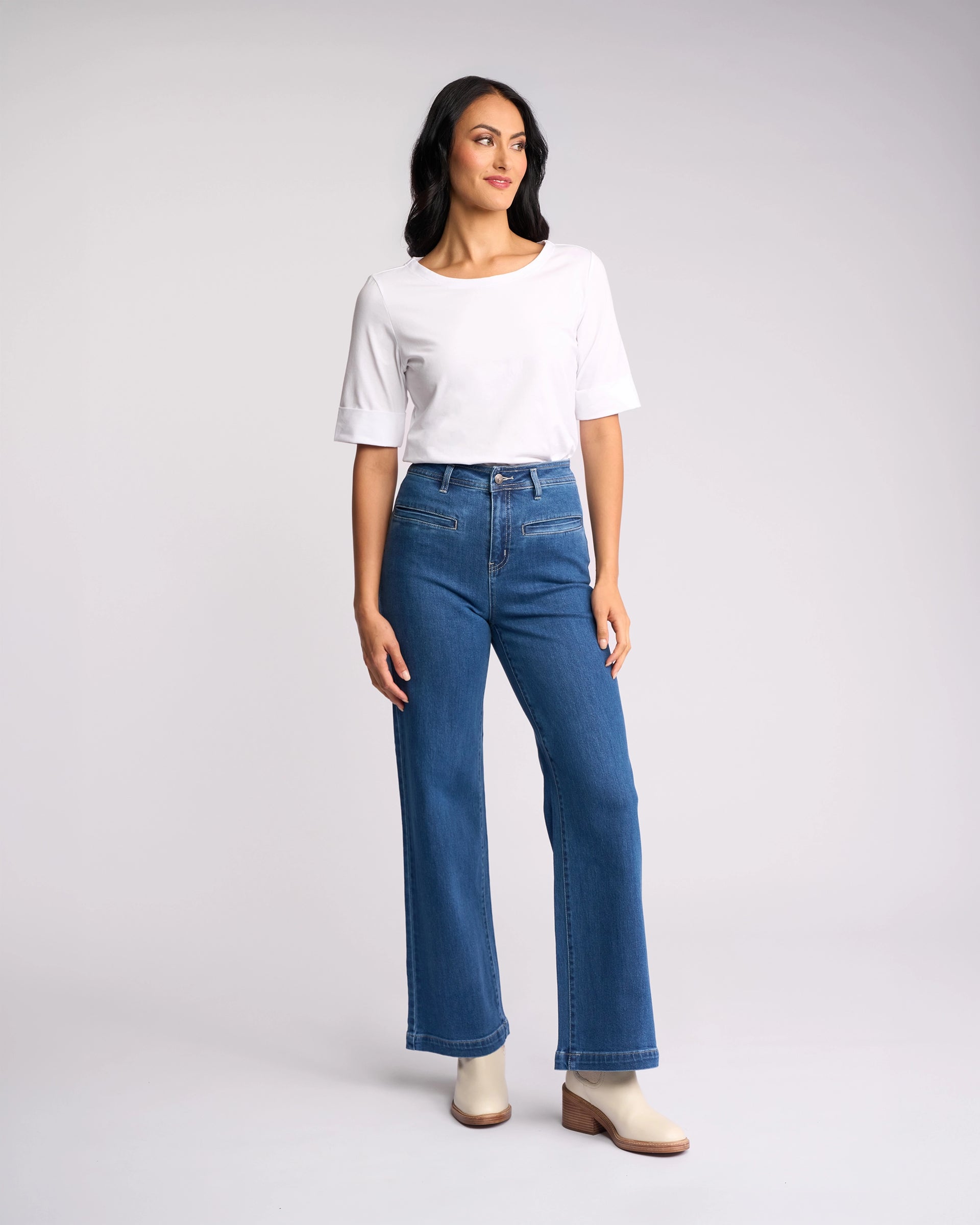 A woman stands against a plain light background, wearing a white short-sleeve top, Vassalli's Peace Jean in blue wash denim, and light-colored ankle boots. She looks to the side with a relaxed posture.