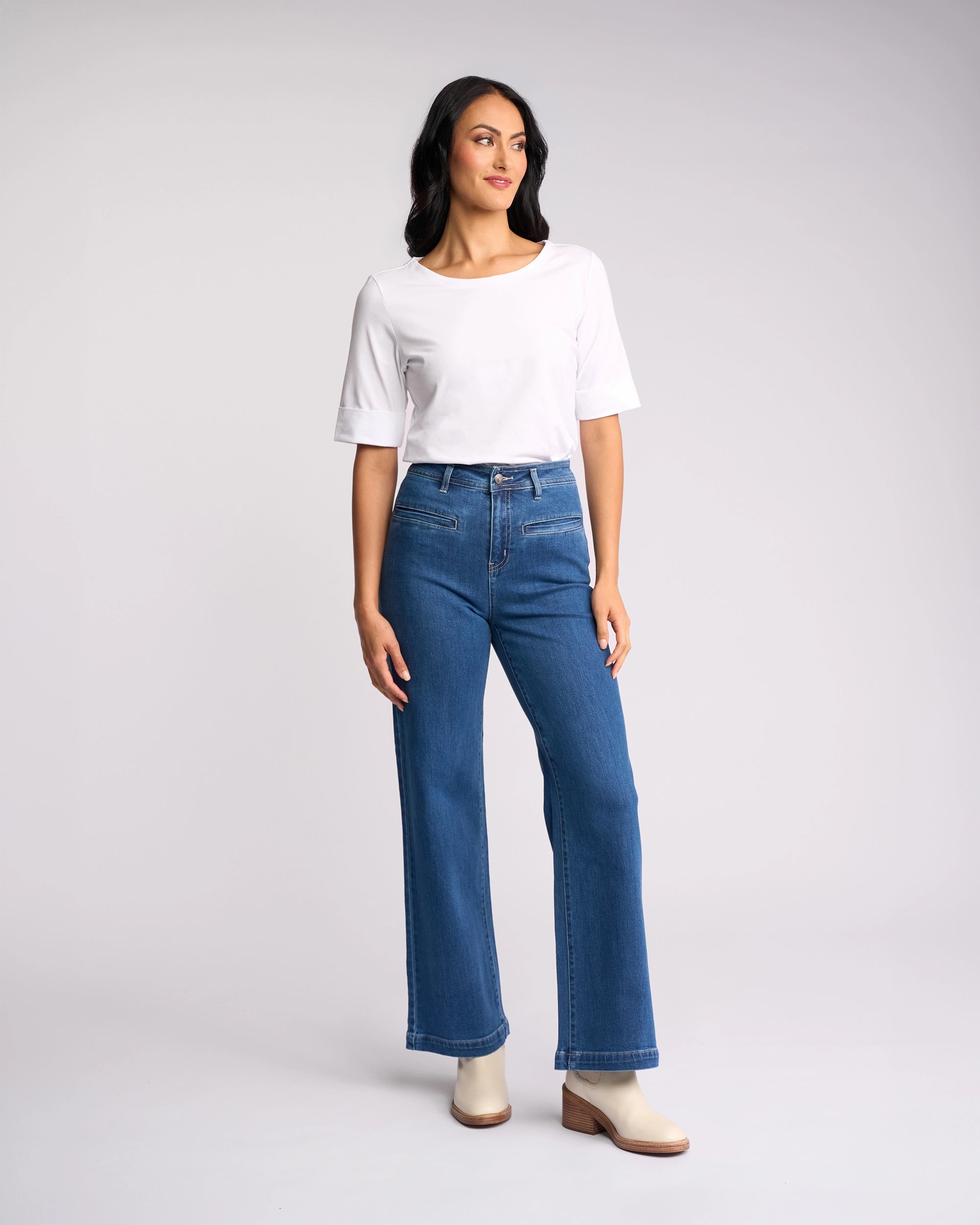 A woman stands against a plain light background, wearing a white short-sleeve top, Vassalli's Peace Jean in blue wash denim, and light-colored ankle boots. She looks to the side with a relaxed posture.