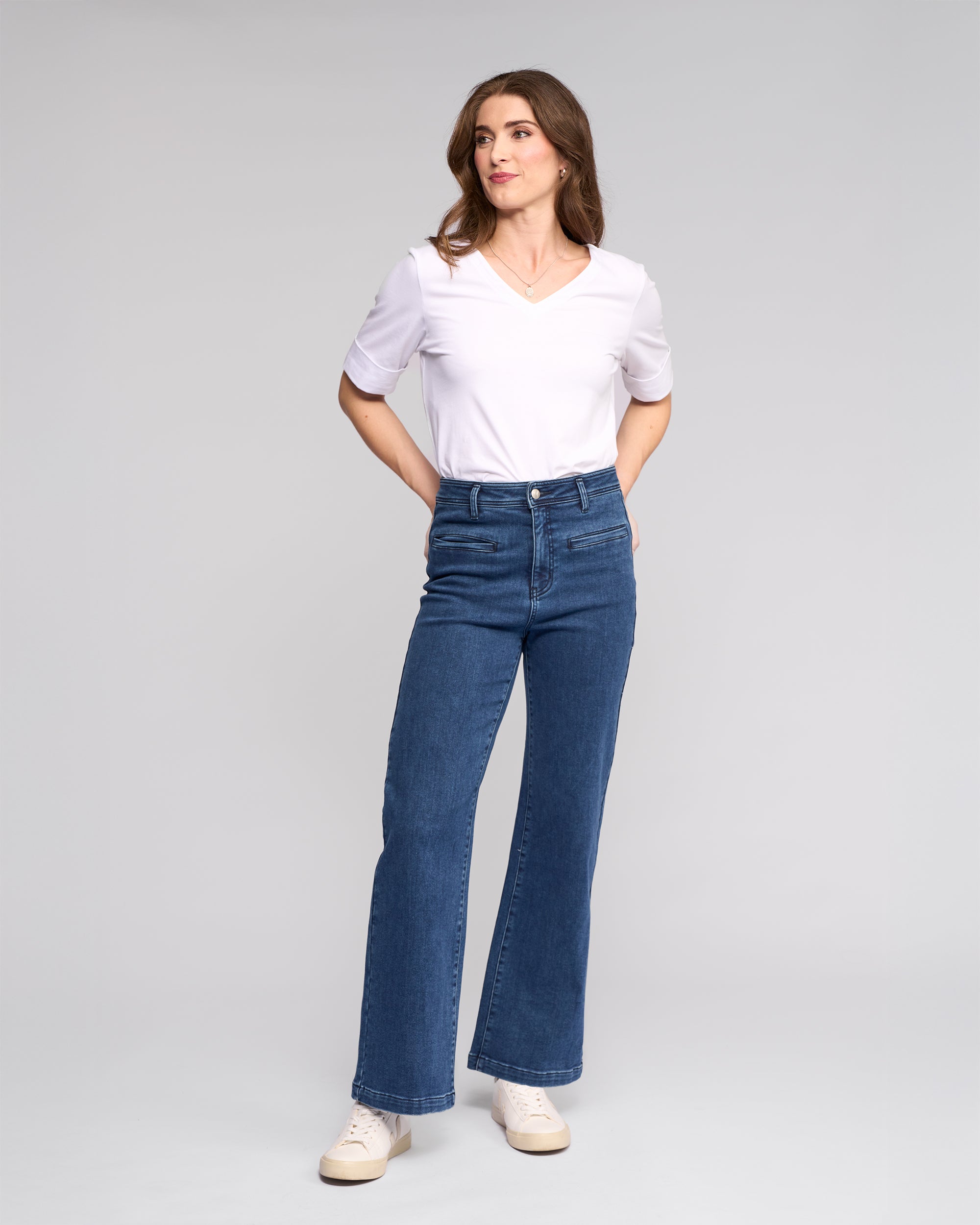 A woman with long brown hair stands against a gray background, wearing a white V-neck T-shirt, Vassalli Peace Jean - Brushed Indigo, and white sneakers, her hands in her pockets as she looks to the side.