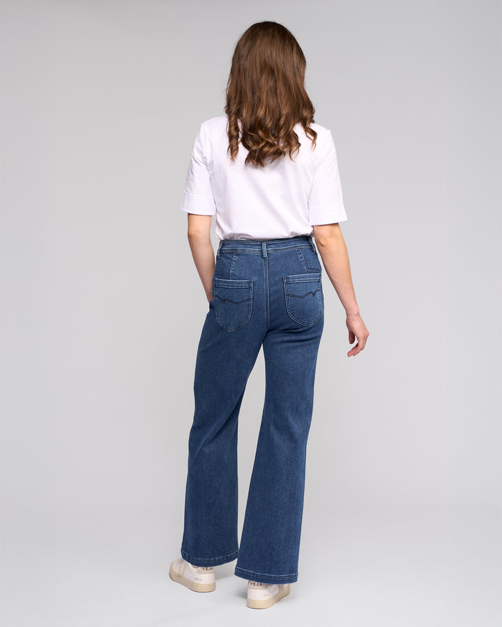 A person with wavy brown hair, dressed in a white short-sleeve shirt and Vassalli Peace Jean - Brushed Indigo, paired with white shoes, stands facing away against a plain light background.