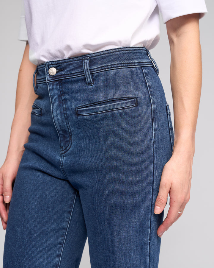 Wearing Vassalli’s Peace Jean in Brushed Indigo, this person sports high-waisted, wide-leg denim with a white short-sleeve shirt, their hand by their side and the look pictured from the torso down for a relaxed silhouette.