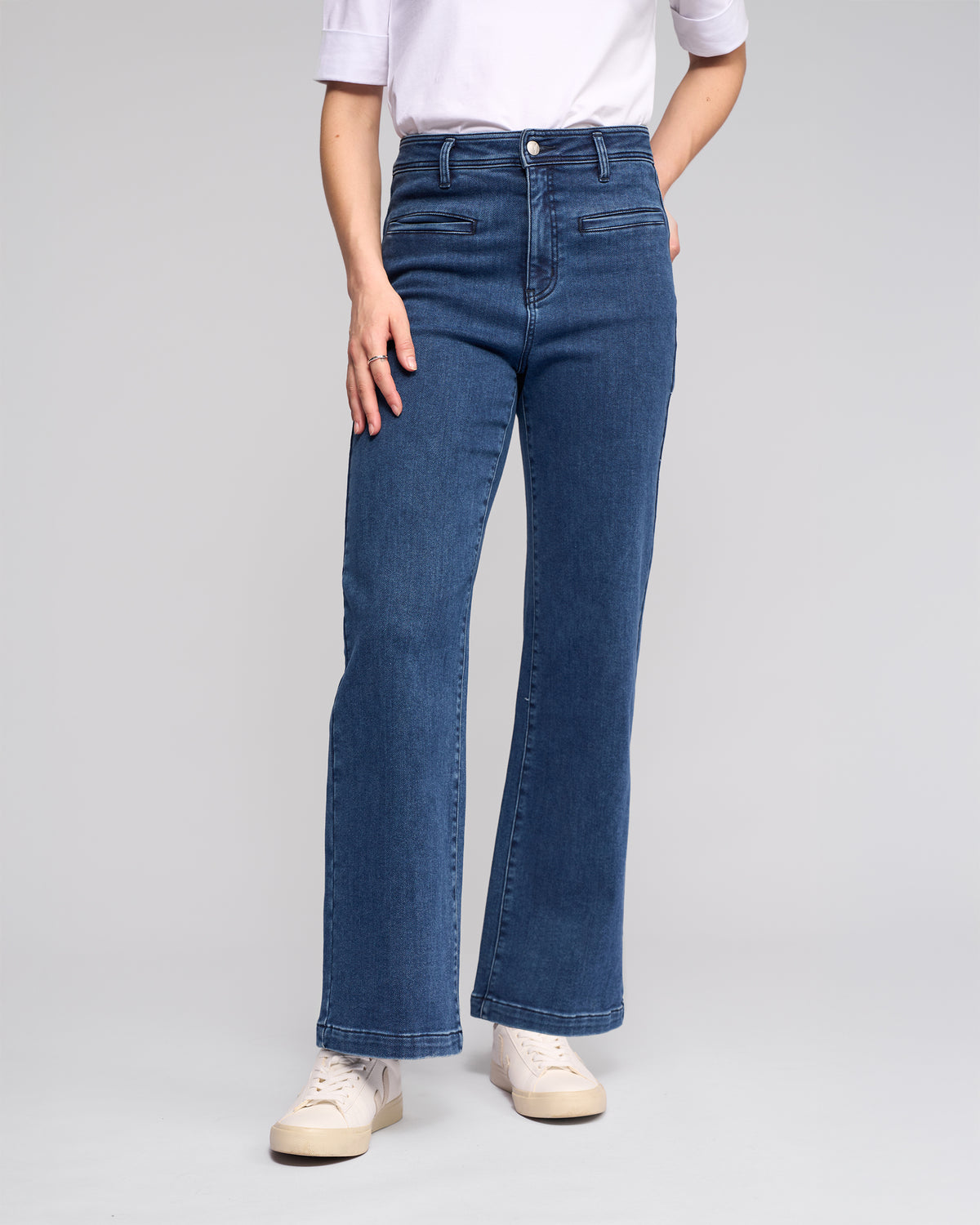 A person wears the Vassalli Peace Jean - Brushed Indigo, paired with a white elbow-sleeve shirt and white sneakers, standing against a plain light gray background. The face is not visible.