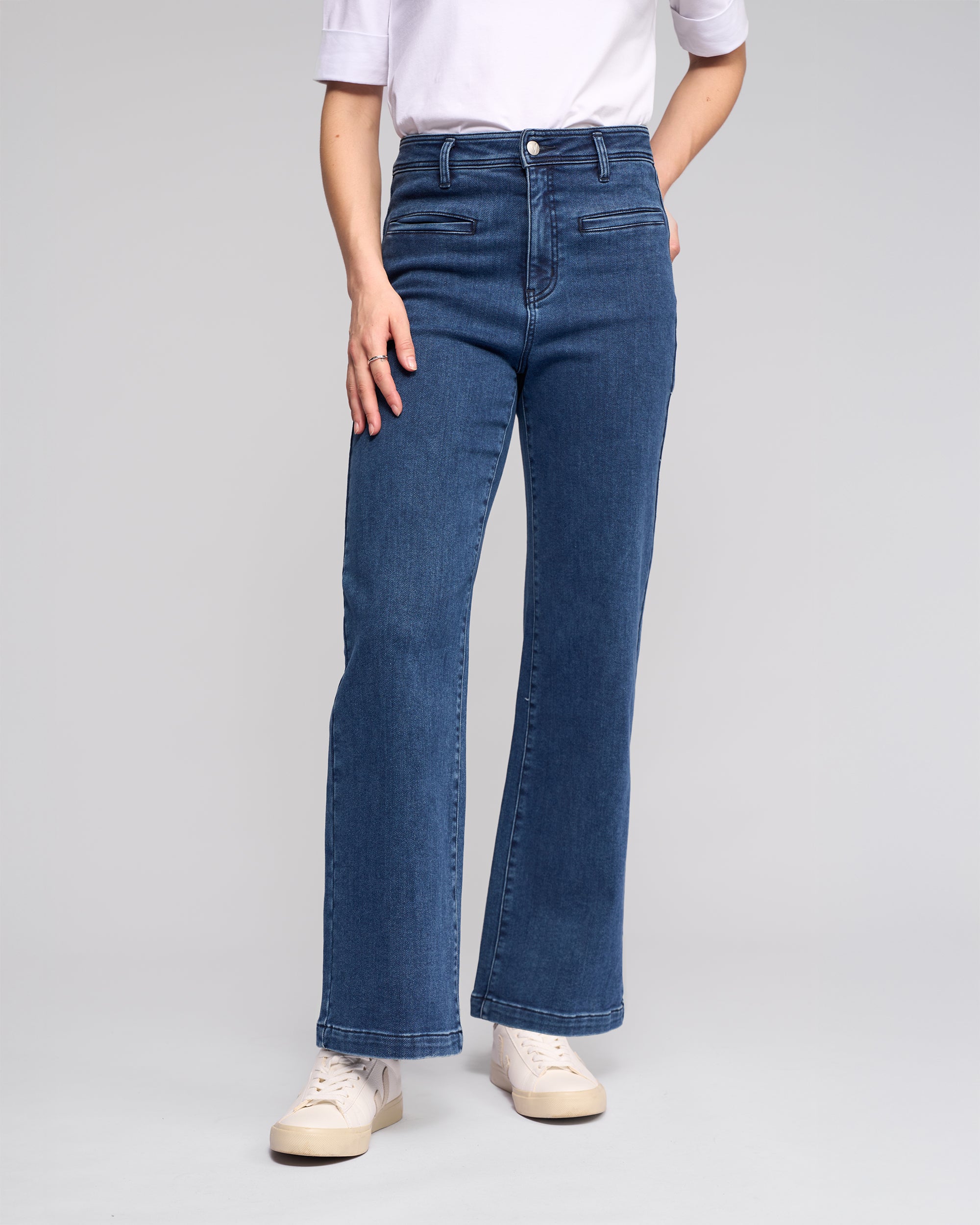 A person wears the Vassalli Peace Jean - Brushed Indigo, paired with a white elbow-sleeve shirt and white sneakers, standing against a plain light gray background. The face is not visible.