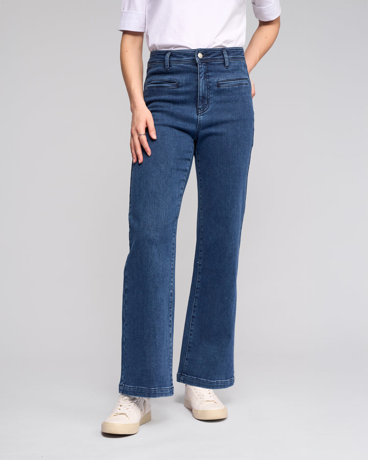 A person wears the Vassalli Peace Jean - Brushed Indigo, paired with a white elbow-sleeve shirt and white sneakers, standing against a plain light gray background. The face is not visible.