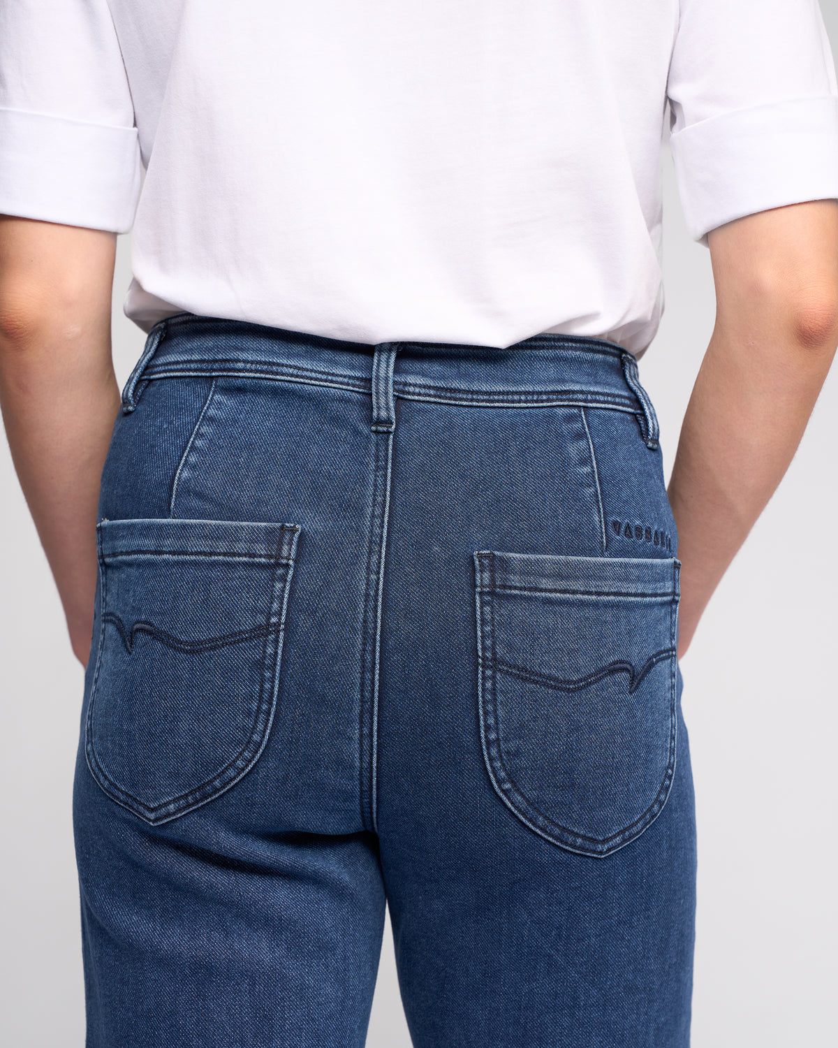Model wears Vassalli Peace Jean - Brushed Indigo, a high-waisted wide-leg denim jean with a relaxed fit, two large back pockets, and decorative stitching, paired with a white short-sleeve shirt. Shown from the back.